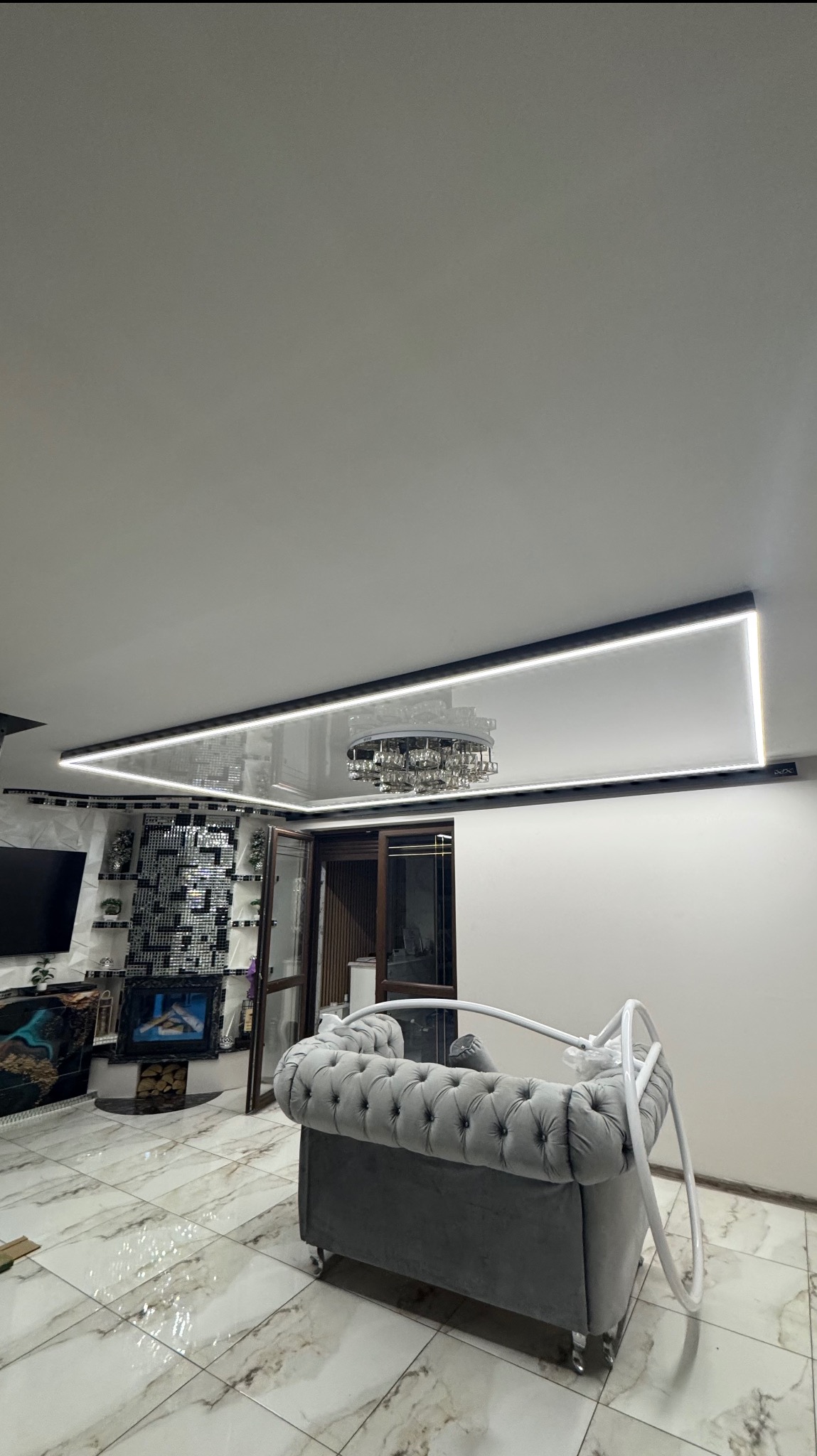 Nowoczesny salon z błyszczącym sufitem podwieszanym, oświetleniem LED i kryształowym żyrandolem. Widoczny szary narożnik i marmurowa podłoga.