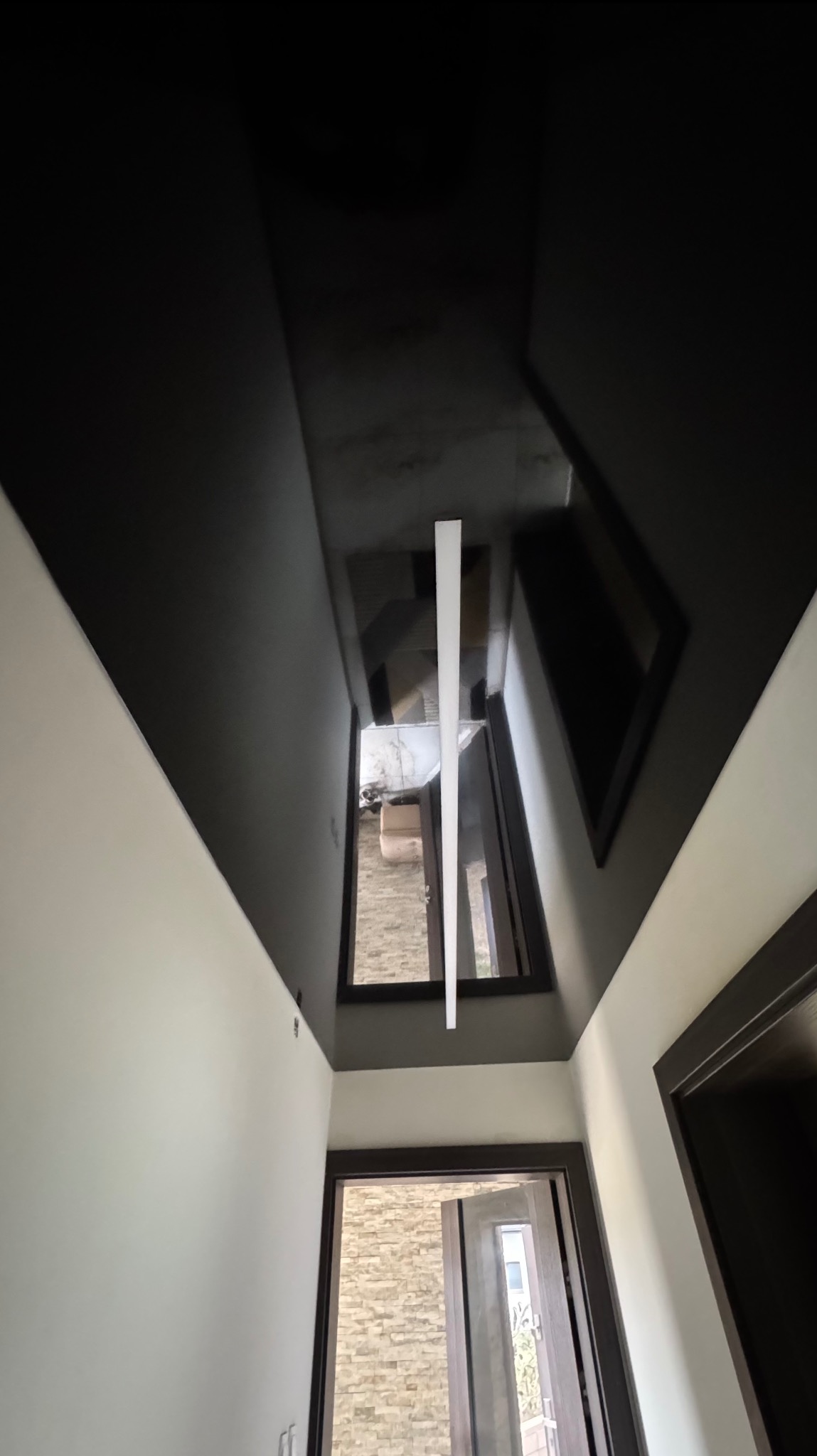 Błyszczący, czarny sufit z oknem dachowym i podłużną lampą LED w nowoczesnym korytarzu z białymi ścianami. Widok z dołu.
