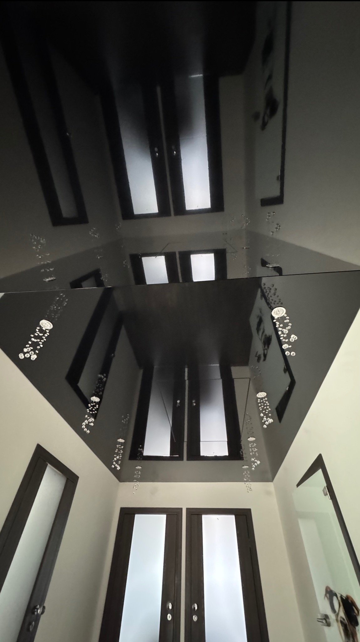 Błyszczący, czarny sufit napinany z dekoracyjnymi elementami w kształcie kropli, odbijający wnętrze z drzwiami i oknami. Widok z dołu.