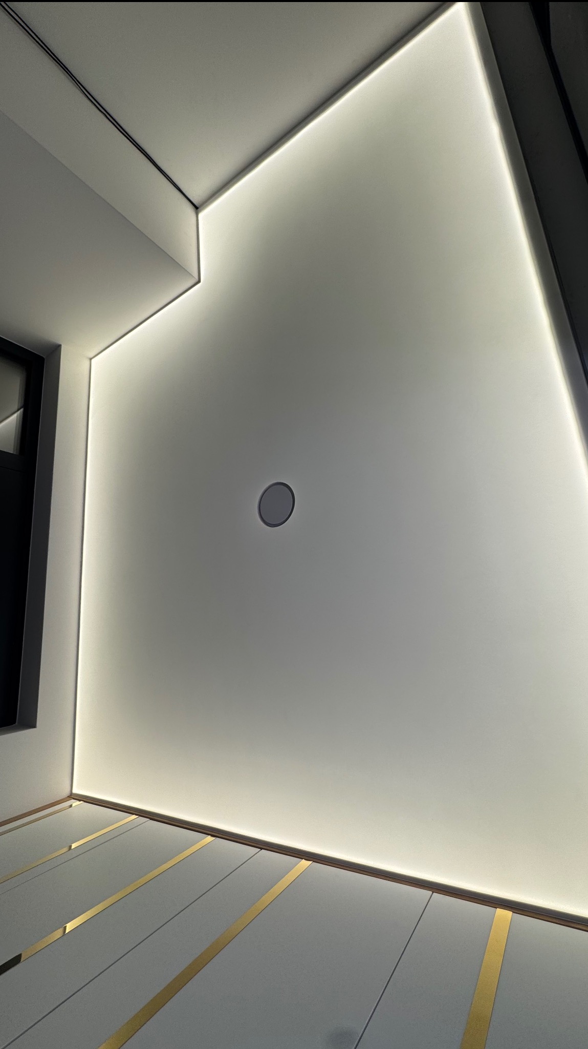 Biały, napinany sufit z oświetleniem LED wzdłuż krawędzi i wbudowanym głośnikiem centralnym. Widoczne panele podłogowe z elementami dekoracyjnymi w kolorze złotym.