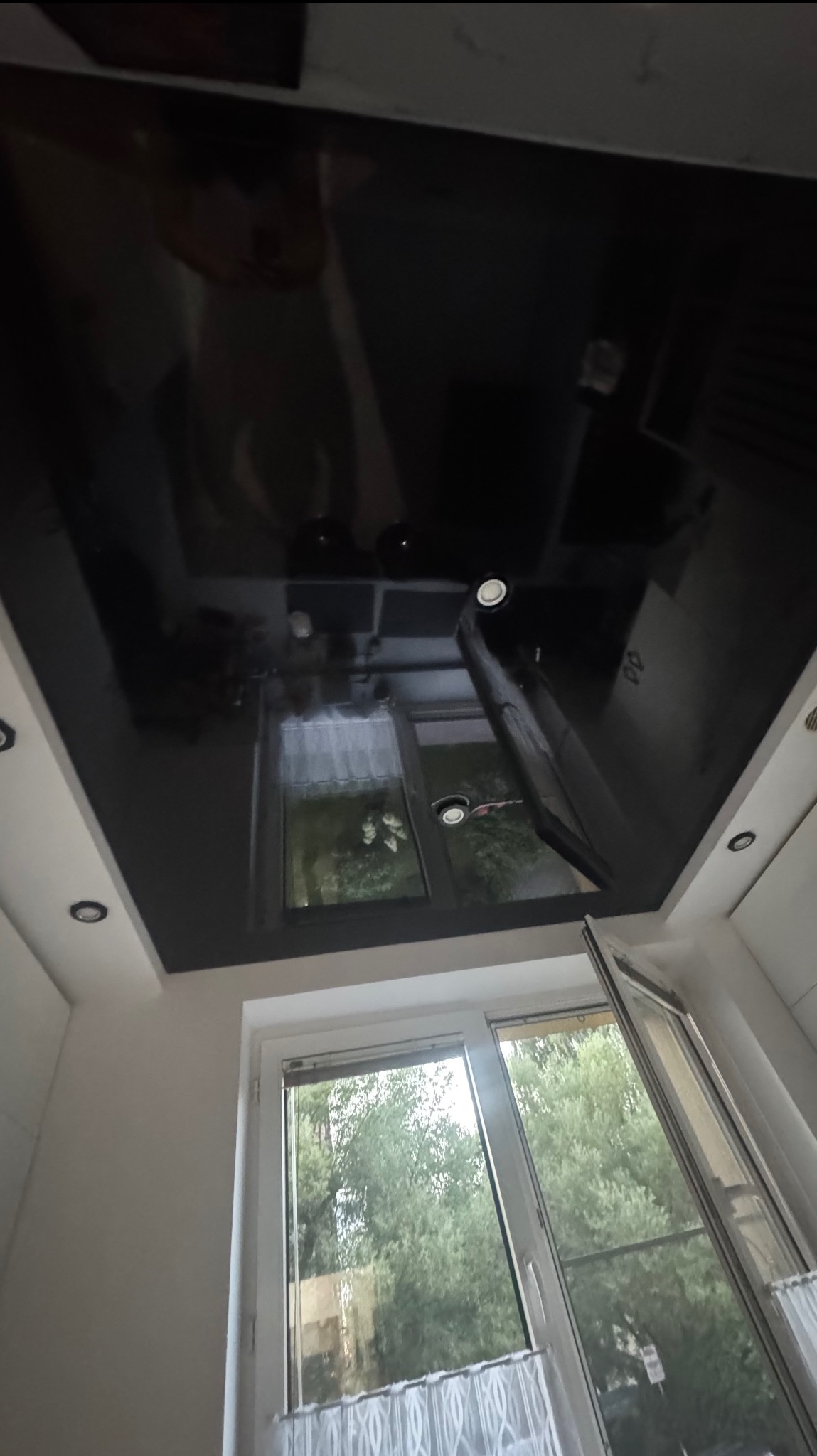 Czarne, lustrzane wykończenie sufitu z wbudowanym oświetleniem, odbijające widok okna z zielenią na zewnątrz. Kąt ujęcia z dołu, widoczne fragmenty ścian.