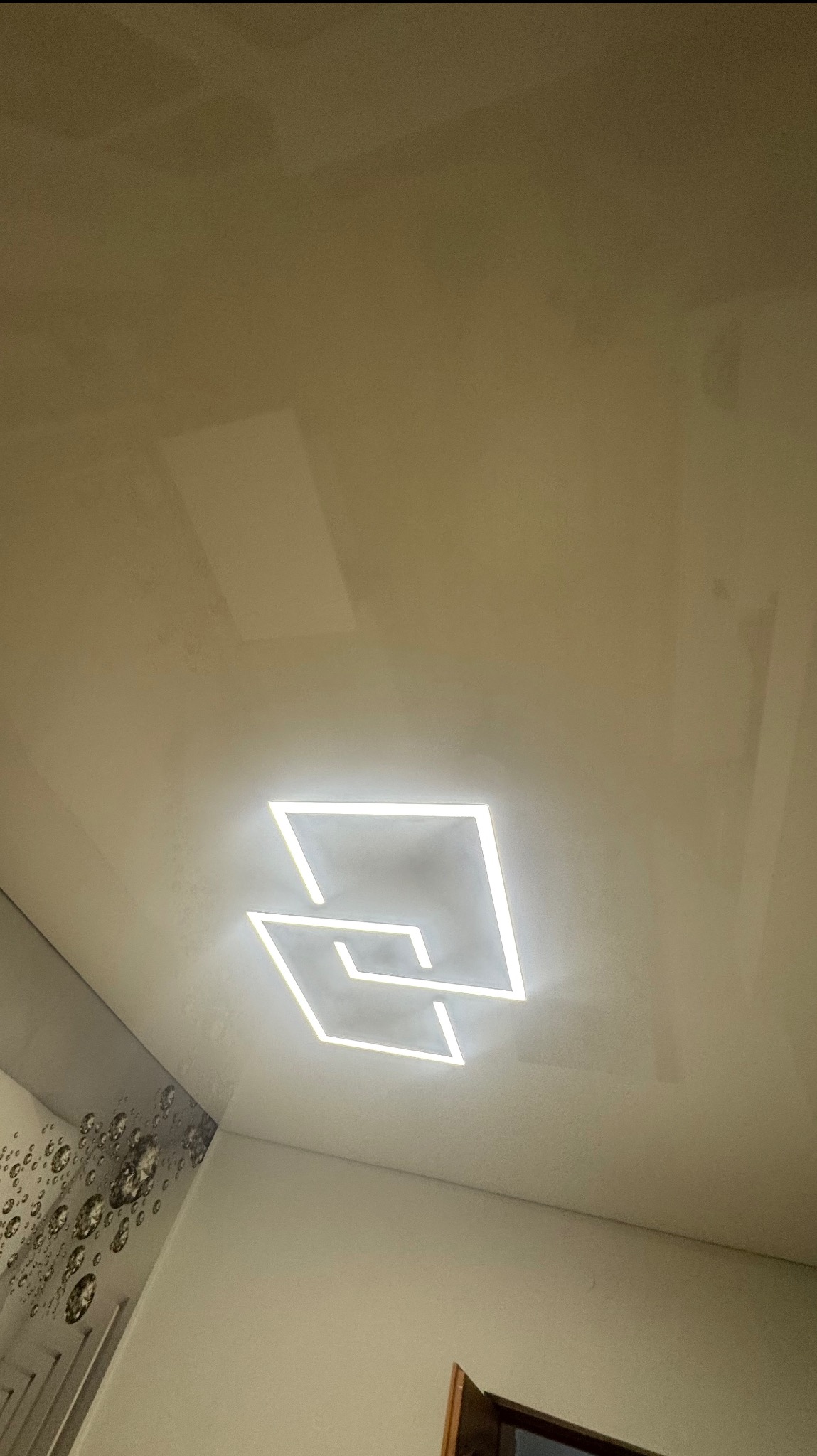 Błyszczący sufit z podwieszonym oświetleniem LED w kształcie dwóch kwadratów, widok z dołu, dekoracyjna ściana w tle.