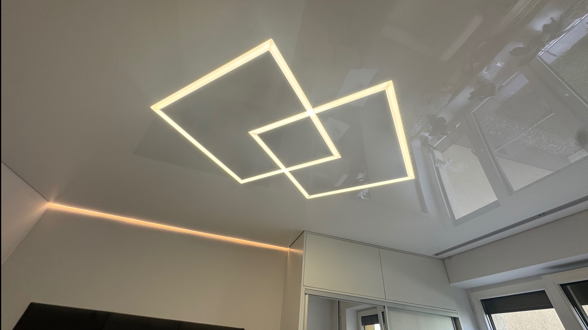 Biały sufit z połyskiem, oświetlenie LED w kształcie dwóch kwadratów, dodatkowe oświetlenie liniowe przy ścianie. Widok na okno i szafę w tle.