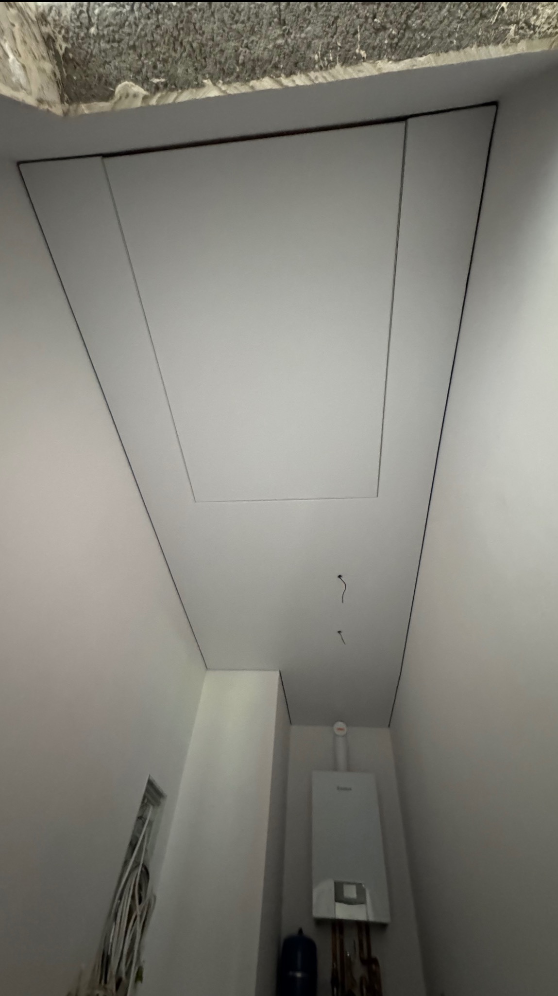 Minimalistyczny, biały sufit z precyzyjnie wkomponowanym włazem rewizyjnym, widoczne przewody i piec gazowy w tle. Kąt ujęcia z dołu podkreśla gładkość powierzchni.