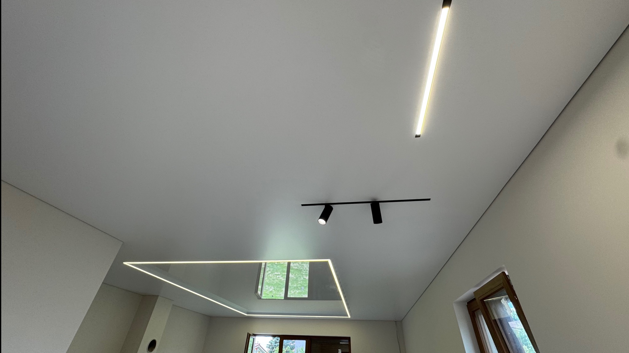 Nowoczesny, biały sufit z oświetleniem LED: podłużna lampa, reflektory na szynie i kwadratowa rama LED wokół okna dachowego.