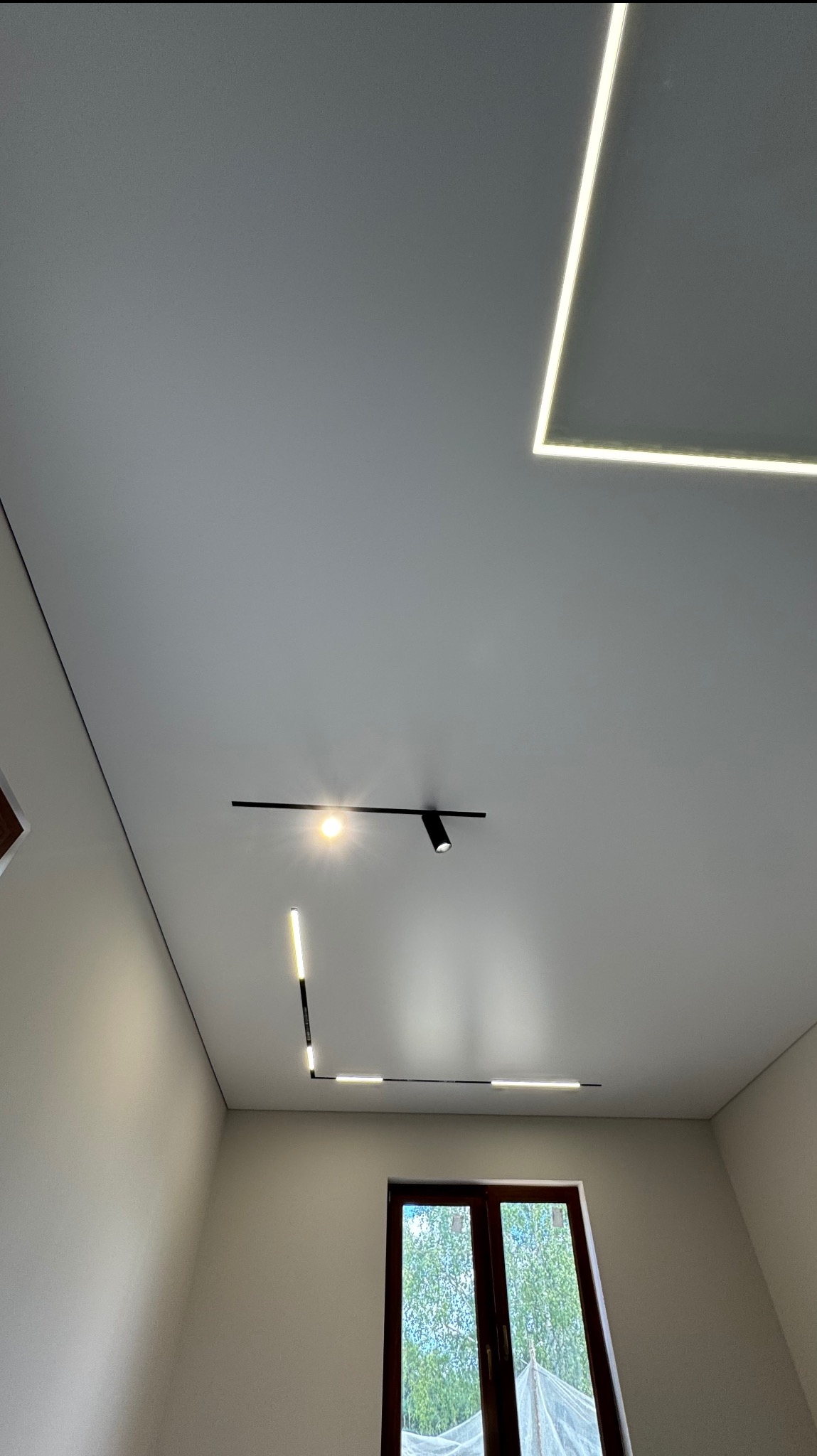 Biały sufit napinany z nowoczesnym oświetleniem LED: kwadratowy panel i liniowe lampy magnetyczne, widok z dołu, okno w tle.