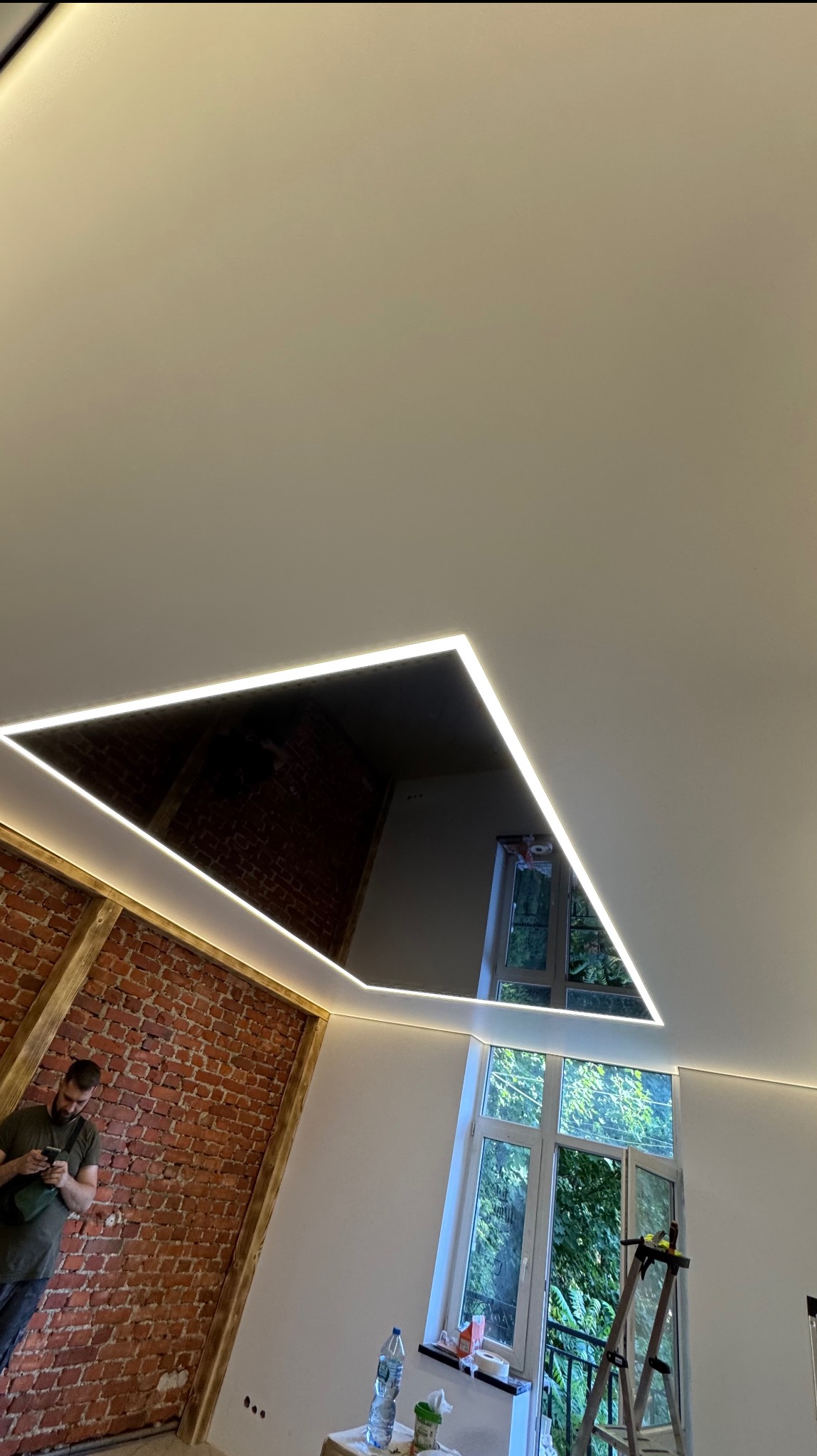 Wykończone wnętrze z lustrzanym, trójkątnym elementem na suficie, podświetlonym listwami LED. Widoczny fragment ceglanej ściany i okno z widokiem na zieleń.