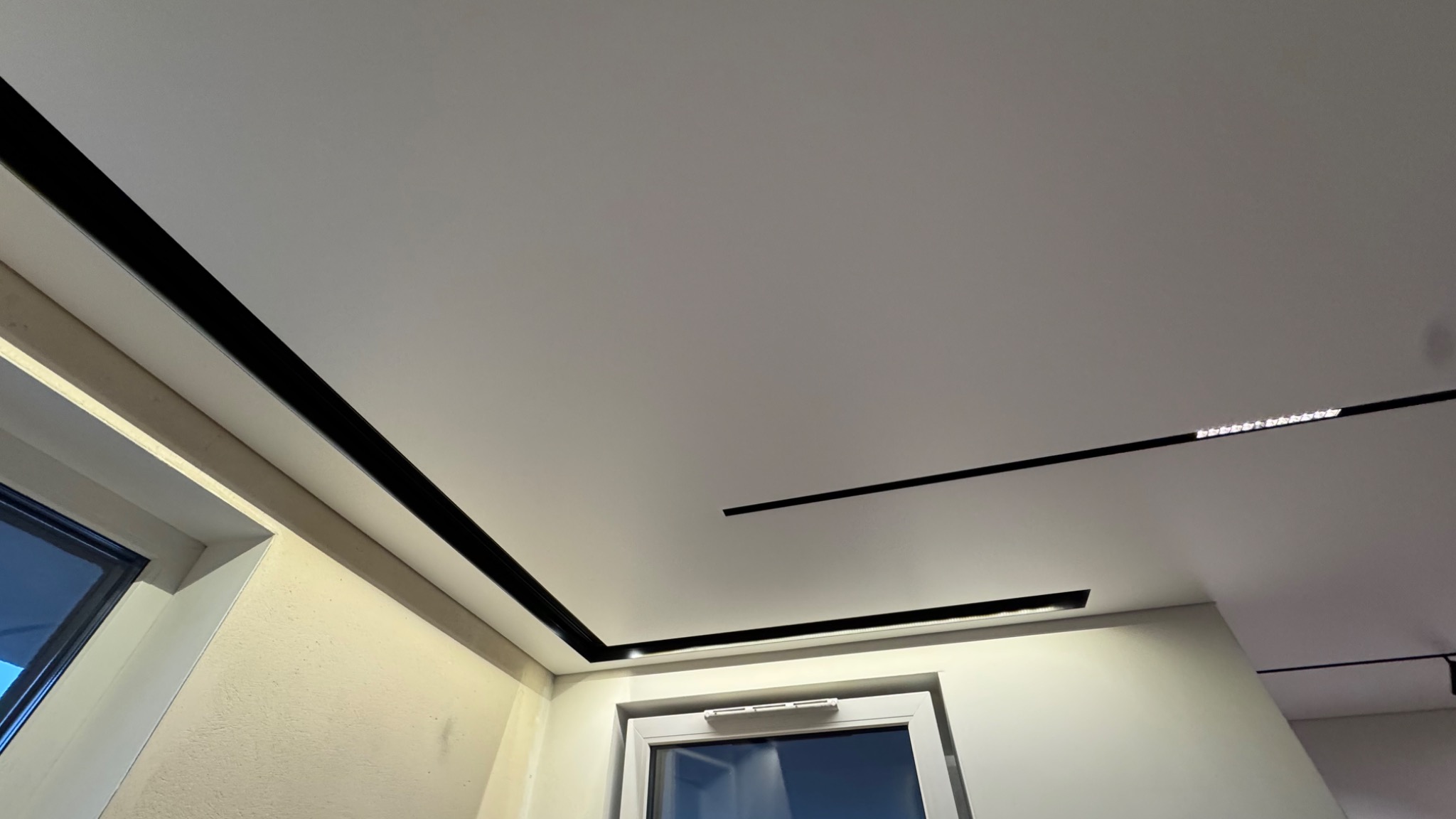 Nowoczesny, biały sufit z czarnymi, minimalistycznymi listwami oświetleniowymi LED, widok z dołu, okno w tle.