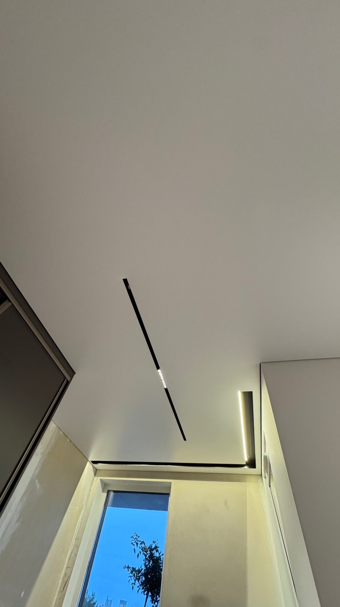 Biały sufit napinany z oświetleniem liniowym LED, widok z dołu, okno z widokiem na zewnątrz. Minimalistyczny design, nowoczesne wykończenie.