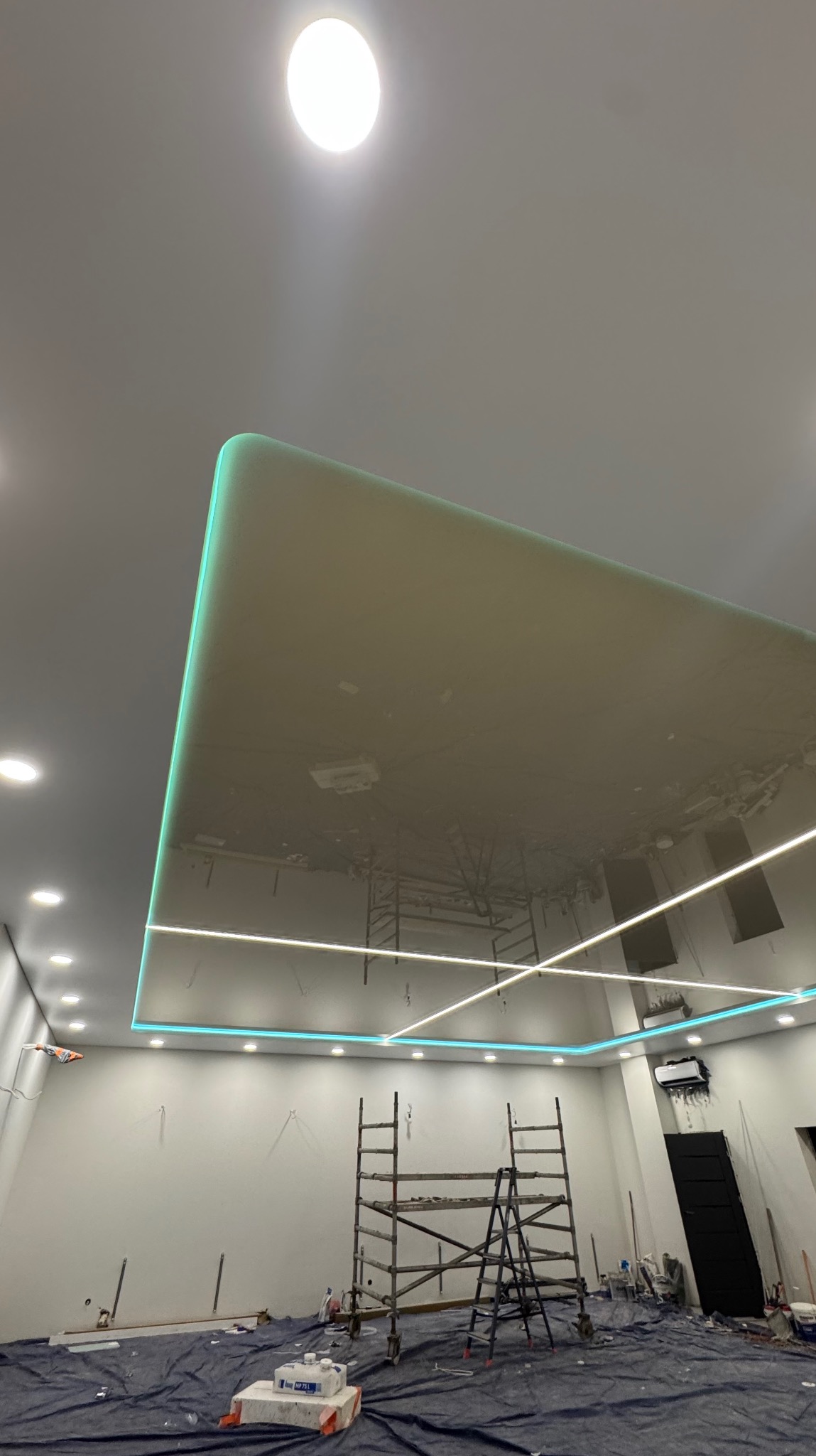 Błyszczący sufit z podświetleniem LED w trakcie montażu, widoczne rusztowania. Białe ściany i zabezpieczona podłoga. Kątowe ujęcie podkreśla geometrię sufitu.