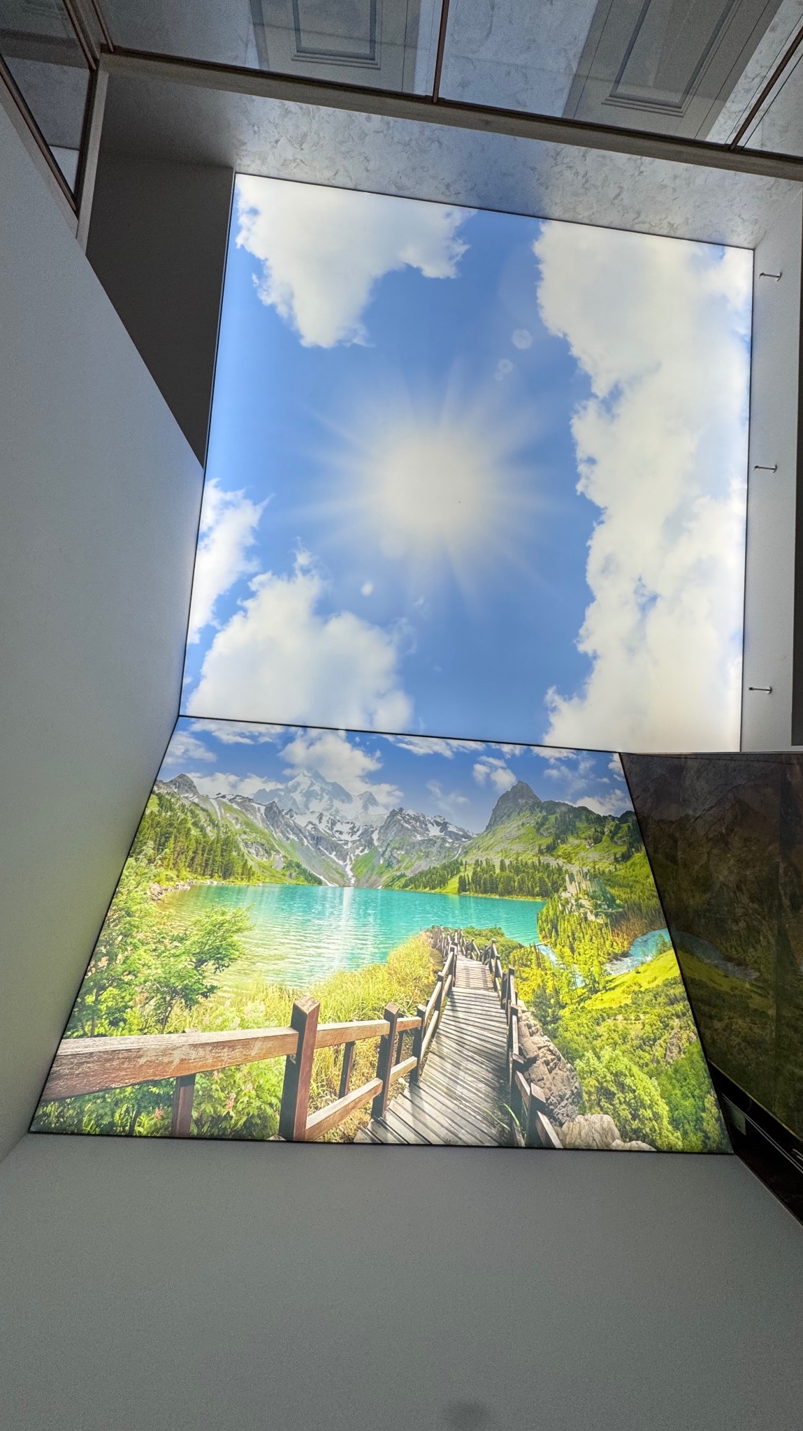 Iluminowany sufit: błękitne niebo z chmurami i słońcem płynnie przechodzi w krajobraz górski z jeziorem i drewnianym mostem. Nietypowa perspektywa.