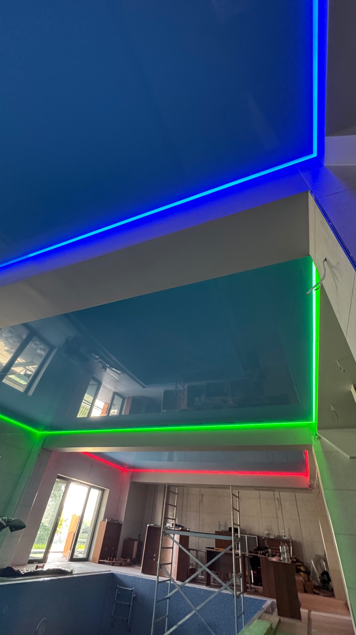 Błękitny sufit z podświetleniem LED w kolorach: niebieskim, zielonym i czerwonym nad basenem. Widoczne rusztowania i okno.