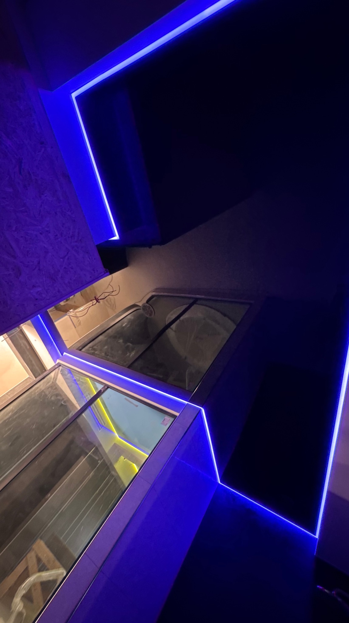 Nowoczesne wnętrze z podwieszanym sufitem i oświetleniem LED w kolorze niebieskim, widziane pod kątem. Widoczne okna dachowe i fragment konstrukcji sufitu.