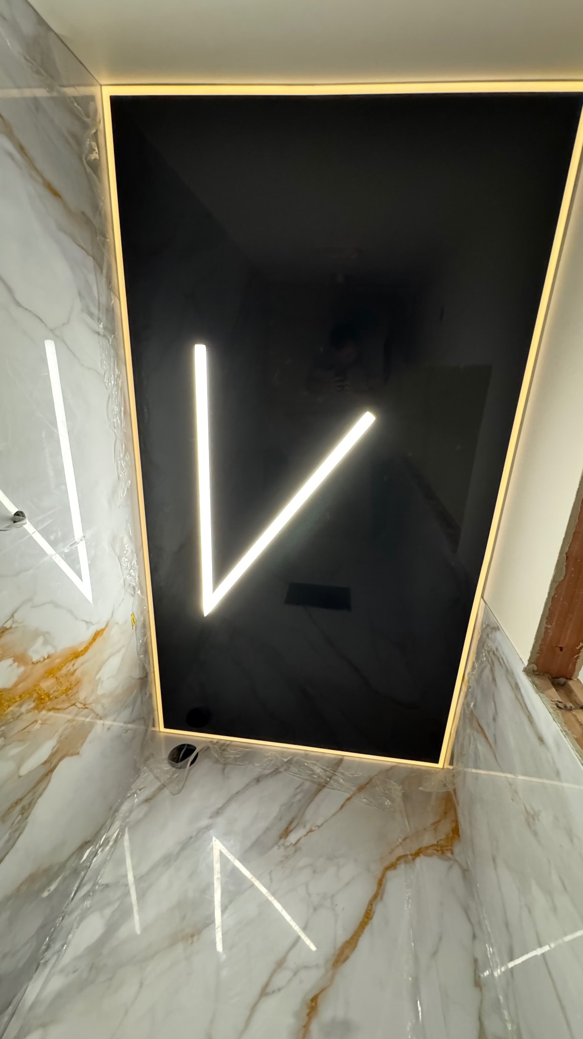 Czarny, błyszczący sufit z podświetleniem LED w kształcie litery V, otoczony złotą ramą. Widoczne marmurowe płytki na ścianach.