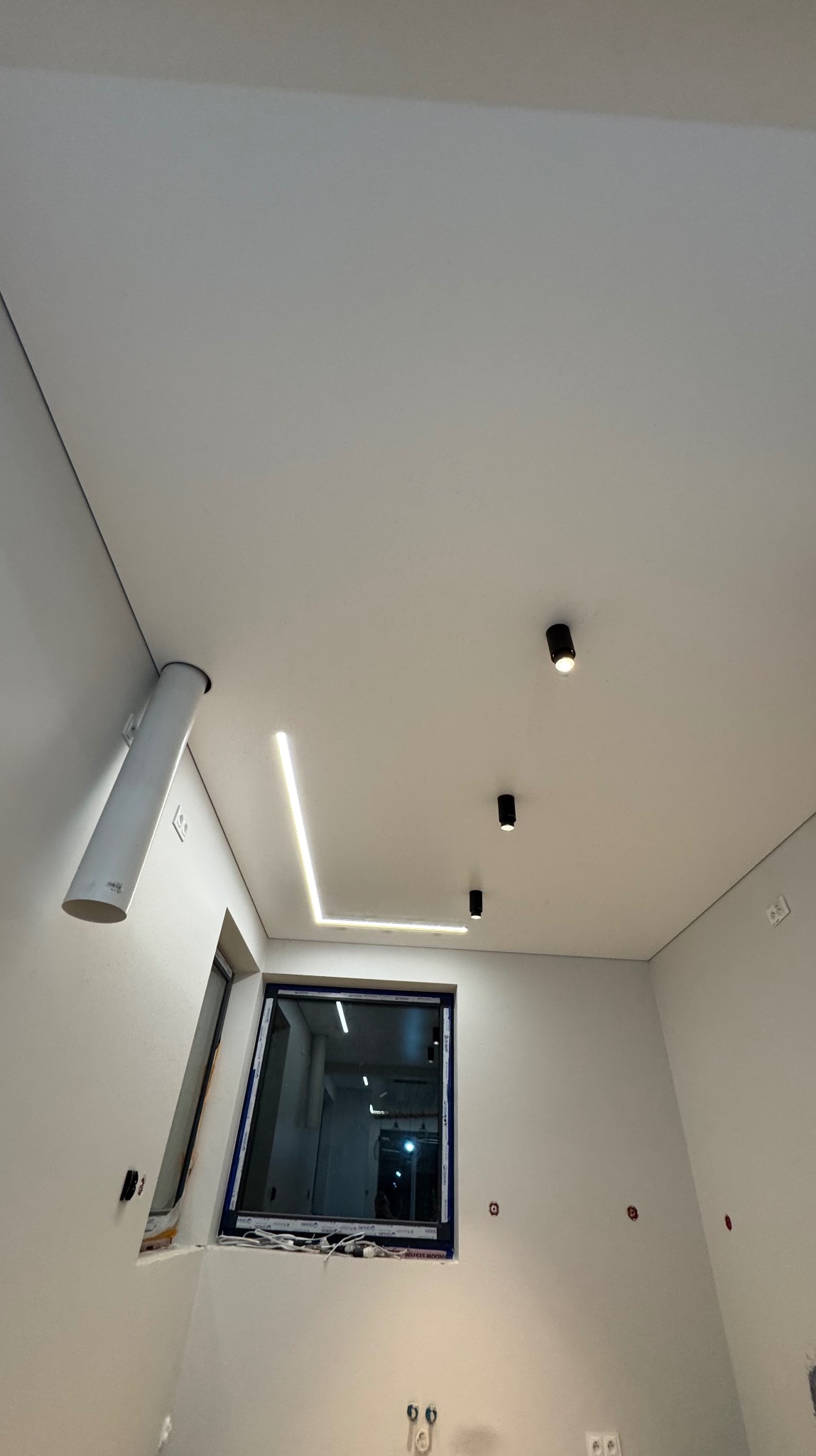 Biały sufit z oświetleniem LED w kształcie litery L i trzema punktowymi lampami. Widoczny fragment instalacji wentylacyjnej i okno w tle. Minimalistyczny design.