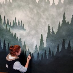 InGraphite Iryna Smorońska - Artystka maluje pędzlem na ścianie leśny motyw z mglistymi drzewami iglastymi. Na podłodze kuweta z farbą i tubki farb akrylowych. Widok z profilu, od pasa w górę.