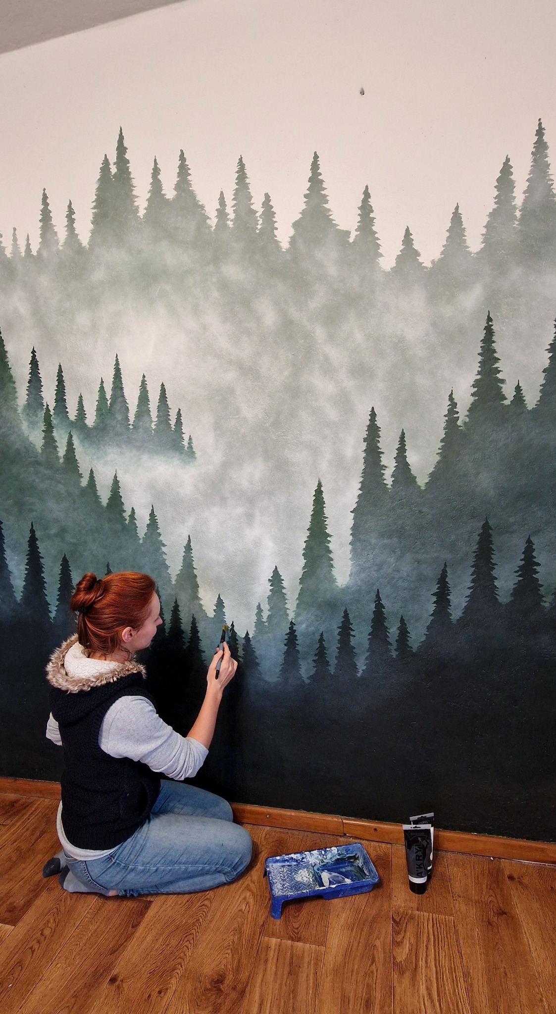 Artystka maluje pędzlem na ścianie leśny motyw z mglistymi drzewami iglastymi. Na podłodze kuweta z farbą i tubki farb akrylowych. Widok z profilu, od pasa w górę.