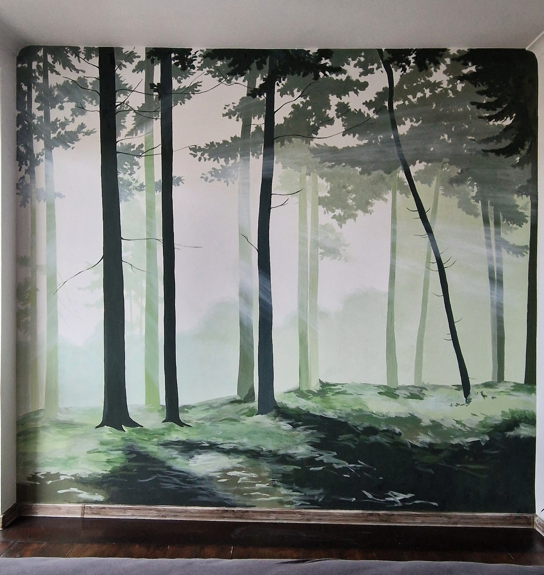 Ściana z malowidłem leśnym, drzewa w odcieniach zieleni i szarości, promienie światła przebijające przez korony. Dekoracja wnętrza w stylu naturalistycznym.