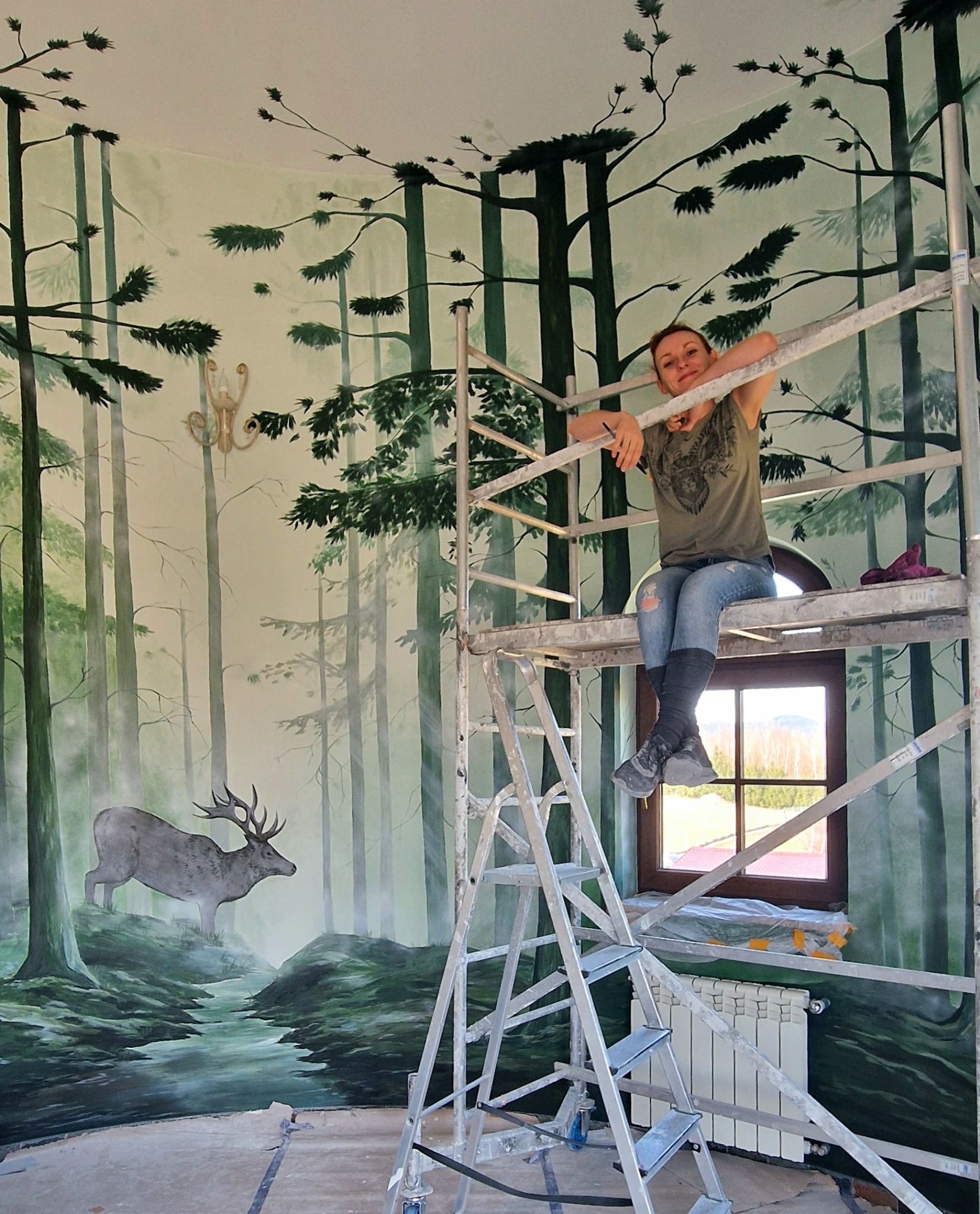 Artystyczne malowanie ścian: kobieta na rusztowaniu, na ścianie las z jeleniem. Widoczne okno i grzejnik, wnętrze w trakcie metamorfozy.