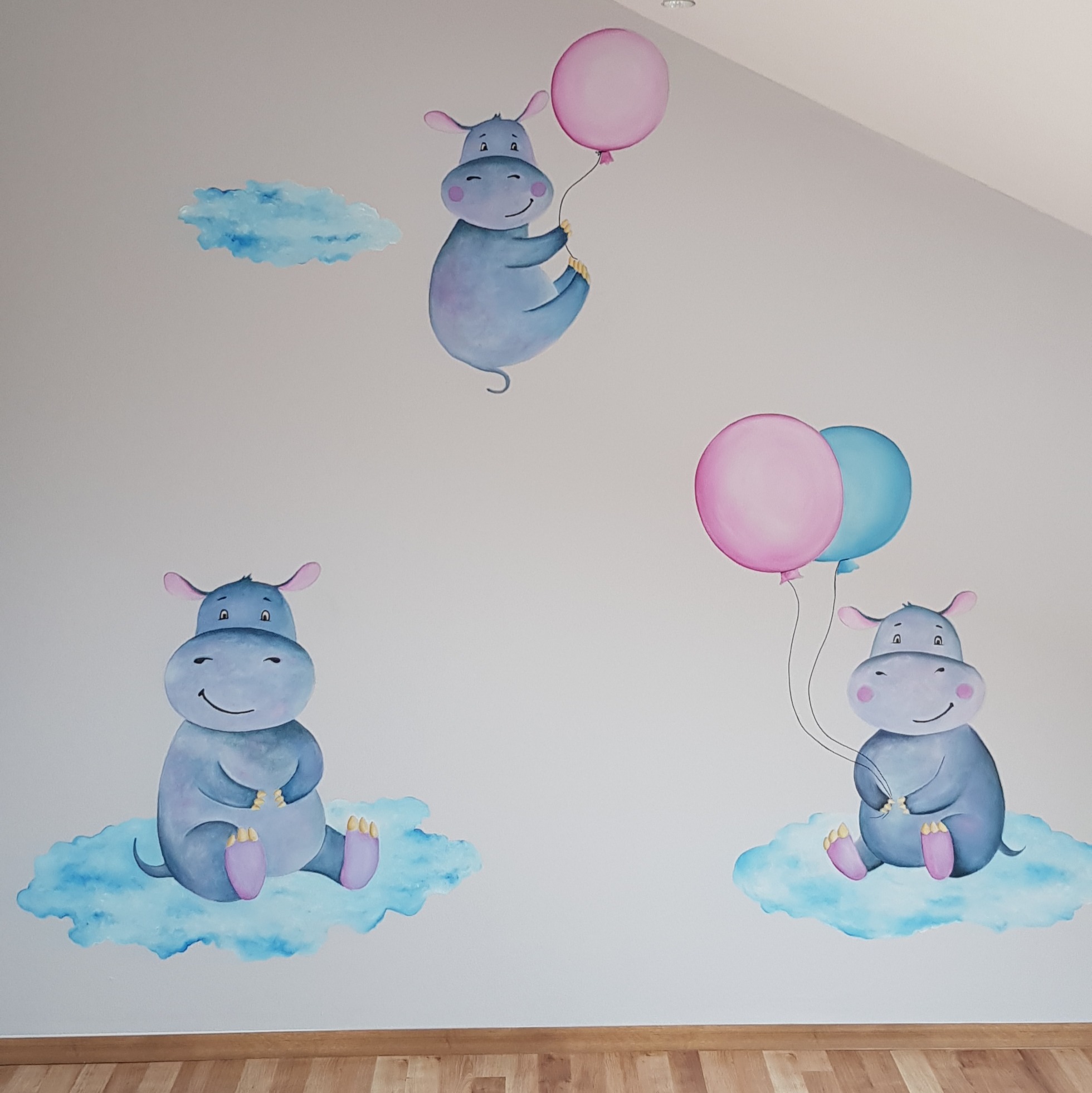 Ręcznie malowana ściana w pokoju dziecięcym: trzy uśmiechnięte hipopotamy na obłokach, jeden z balonem. Jasne kolory, drewniana podłoga, ukośna ściana.