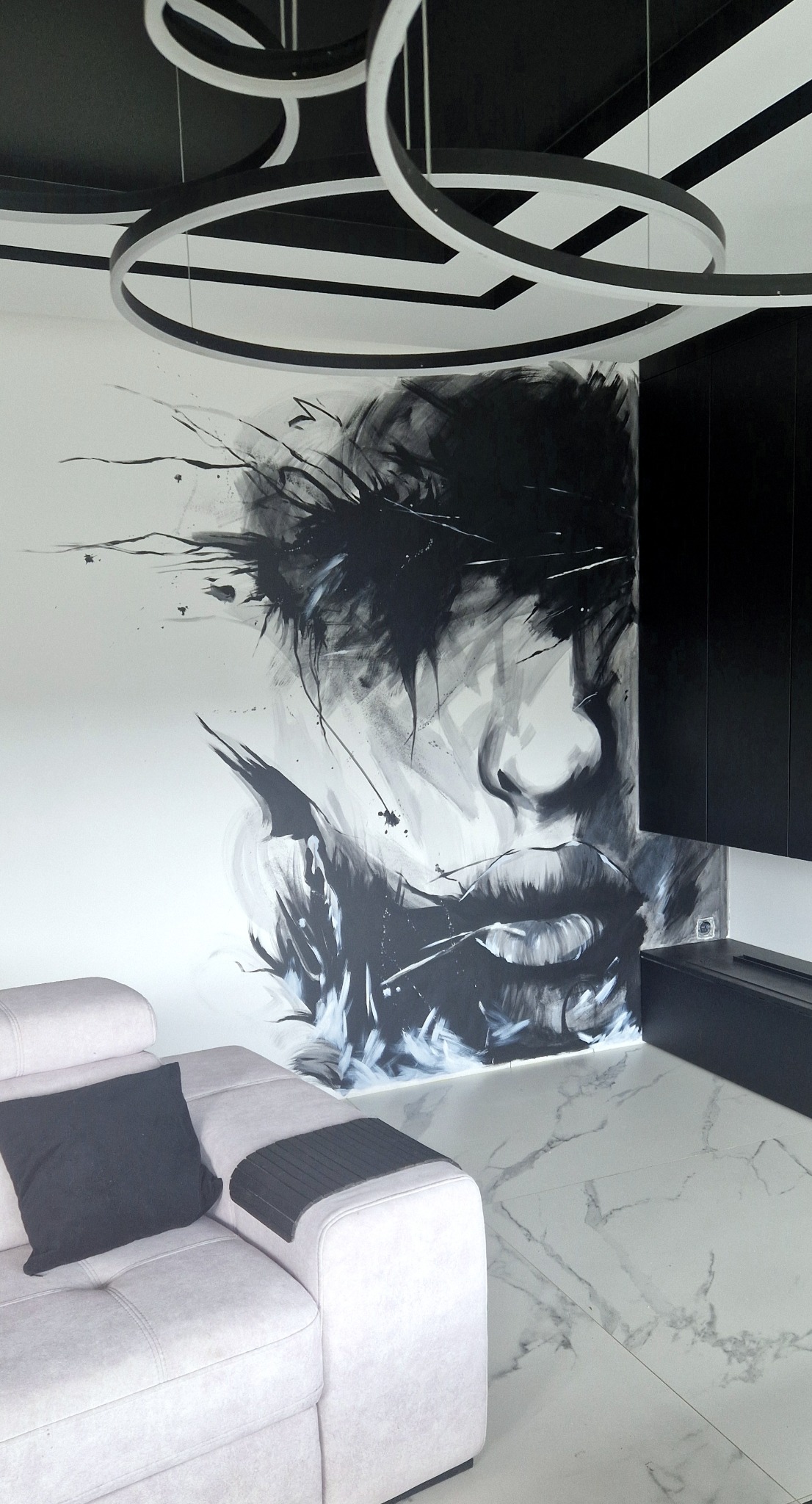 Monochromatyczny mural z abstrakcyjnym portretem na ścianie w nowoczesnym salonie, z elementami oświetlenia i mebli w czerni i bieli.