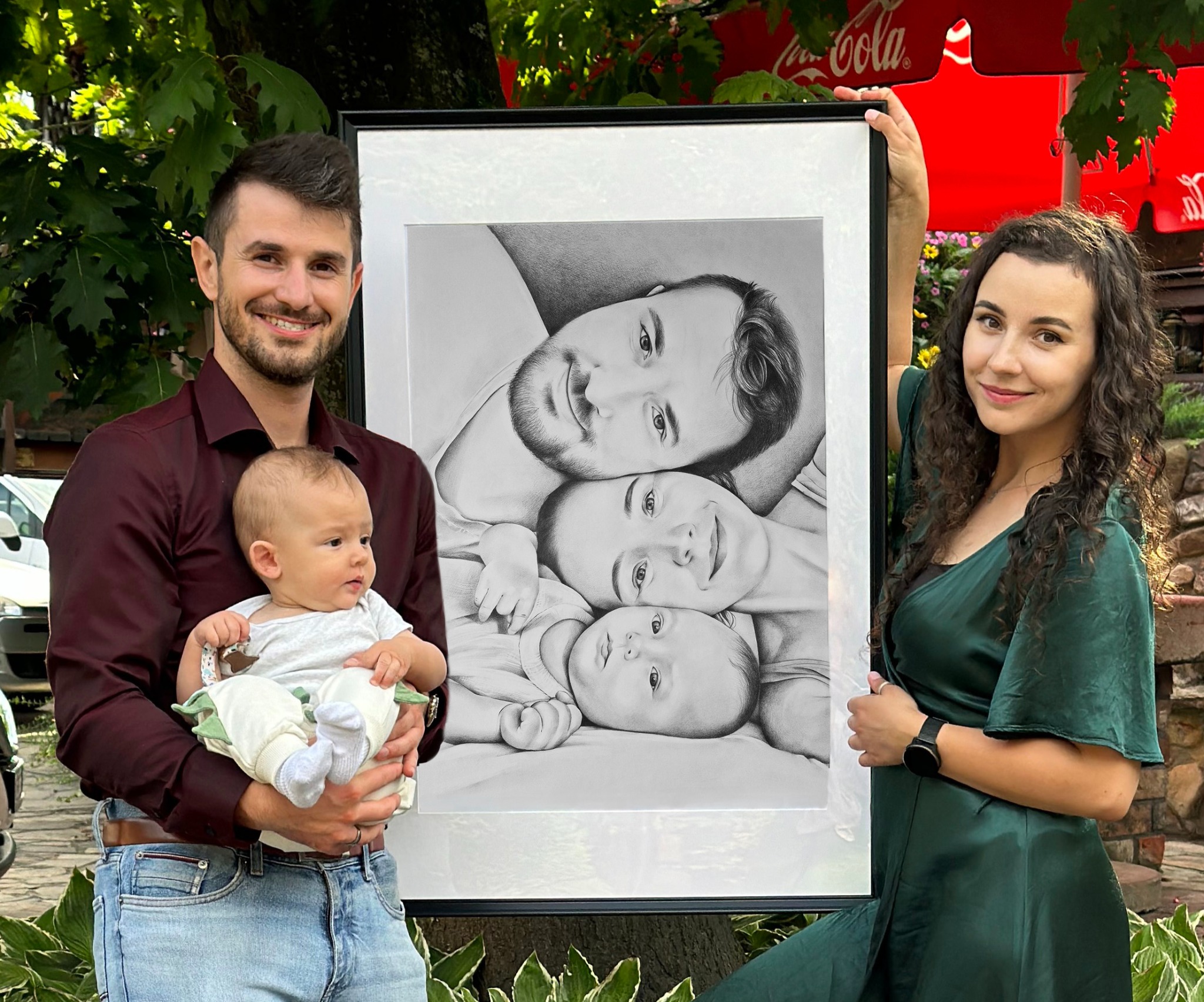 Portret rodzinny 50x70 cm, wykonany ołówkiem, w czarnej ramie, prezentowany przez uśmiechniętych rodziców z dzieckiem na tle zieleni.