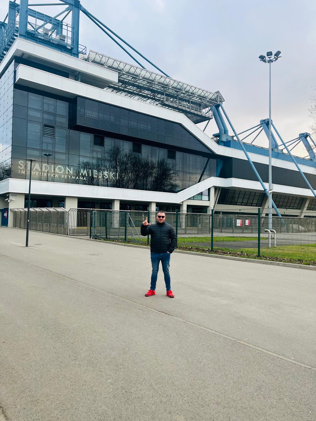 Mężczyzna w kurtce i czerwonych butach stoi przed Stadionem Miejskim im. Henryka Reymana, wskazując palcem do góry. Widoczny fragment fasady i otoczenie stadionu.