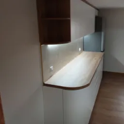 Biała zabudowa kuchenna z drewnianym blatem i półkami, zaokrąglone krawędzie, oświetlenie LED podszafkowe, widok z boku, podłoga drewniana.