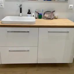 Nowoczesna szafka łazienkowa z białą umywalką i blatem z drewna. Widoczne akcesoria kosmetyczne i fragment kabiny prysznicowej. Minimalistyczny design.