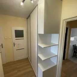 Biała szafa wnękowa z przesuwnymi drzwiami i otwartymi półkami, oświetlona lampą sufitową w korytarzu z drewnianą podłogą i białymi drzwiami.