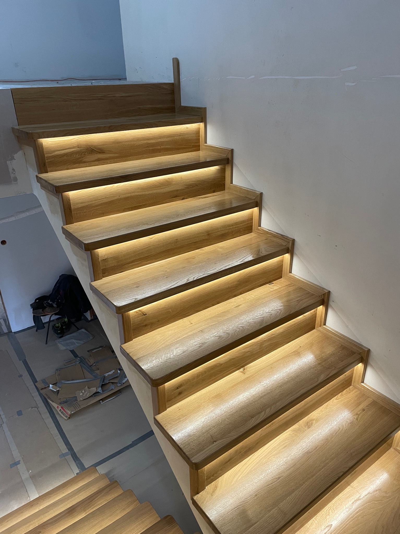 Drewniane schody z subtelnym oświetleniem LED, widoczne z boku, z elementami wykończeniowymi i kartonami w tle, sugerującymi etap montażu.