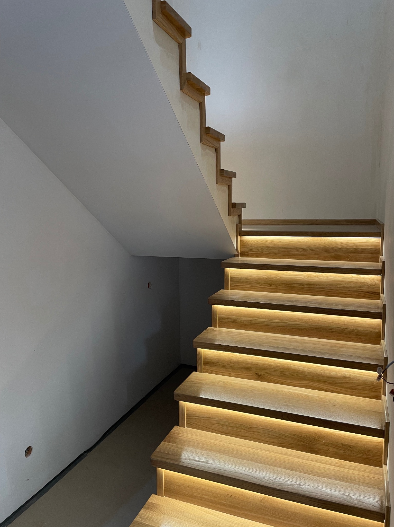 Nowoczesne drewniane schody z podświetleniem LED. Stopnie z jasnego drewna, minimalistyczny design, białe ściany. Widok z dołu, oświetlenie akcentujące.