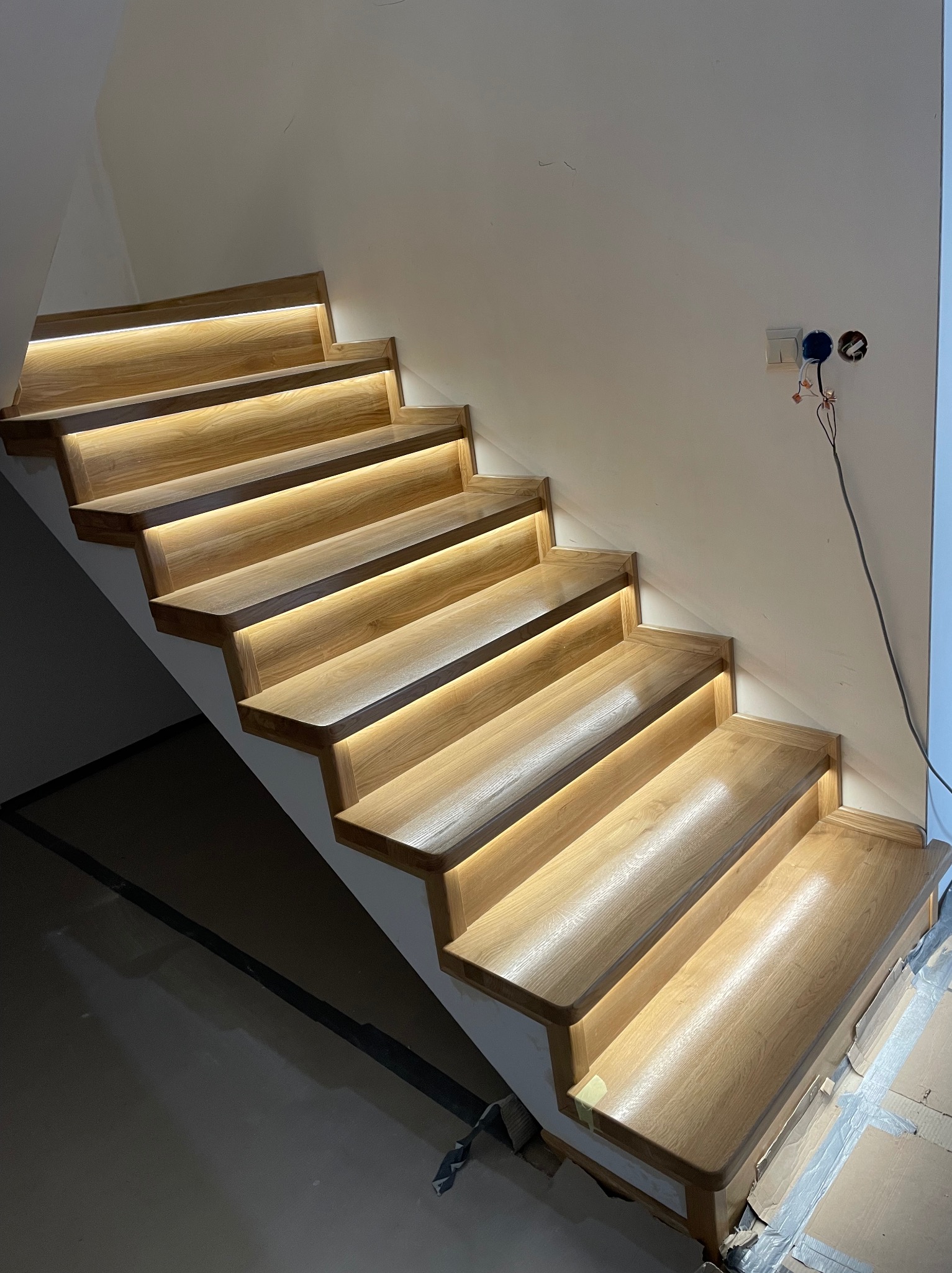 Drewniane schody z oświetleniem LED, widok z dołu. Detal: przewody elektryczne widoczne na ścianie, co sugeruje etap instalacji.