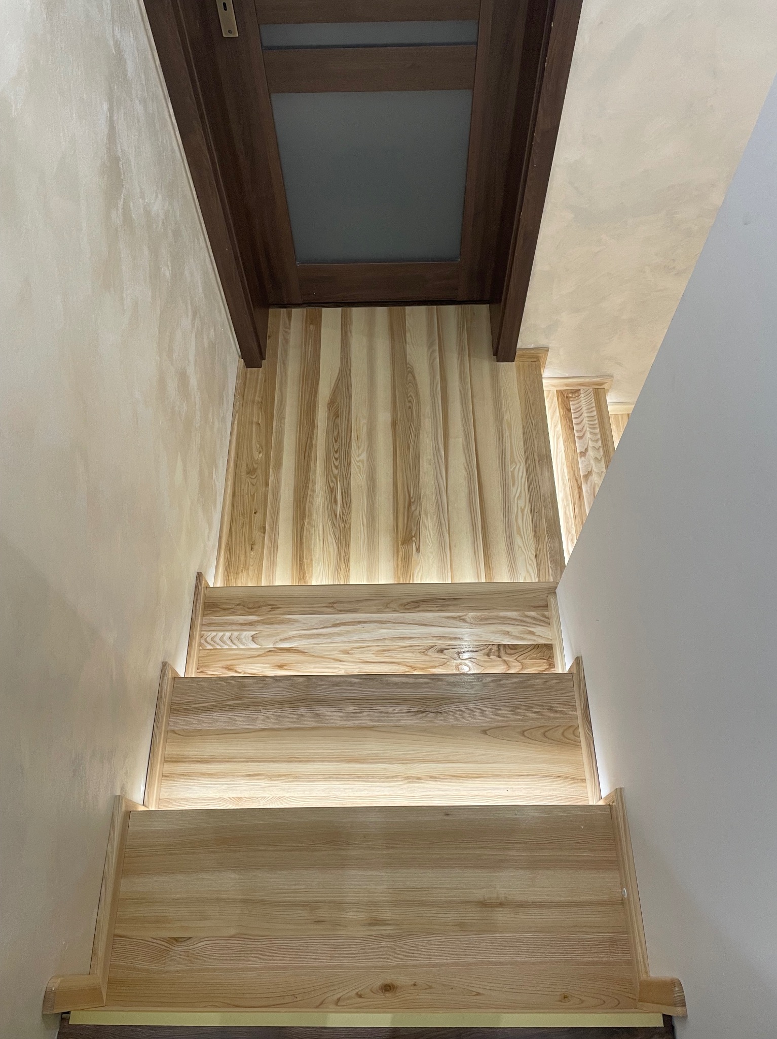 Jasne, drewniane schody z podświetleniem LED, prowadzące w dół, widok z góry. Ściany w odcieniach beżu i bieli, ciemne drzwi na górze schodów.