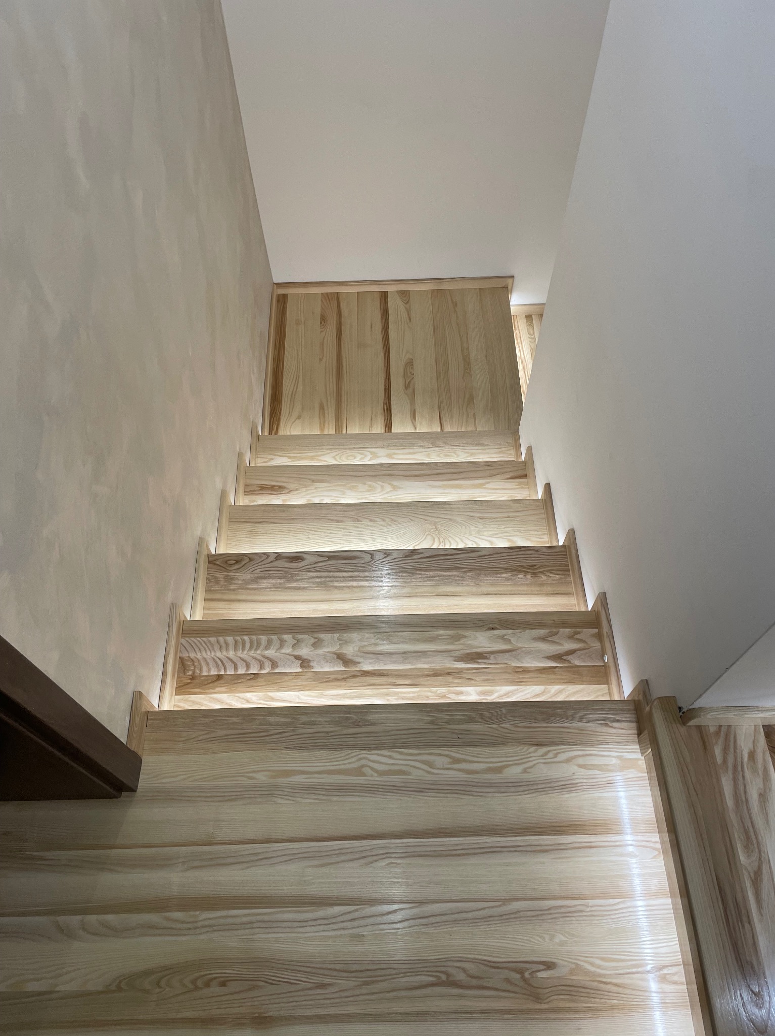 Jasne, drewniane schody z subtelnym podświetleniem LED, prowadzące w górę. Widok z dołu, ukazujący strukturę drewna i minimalistyczny design.