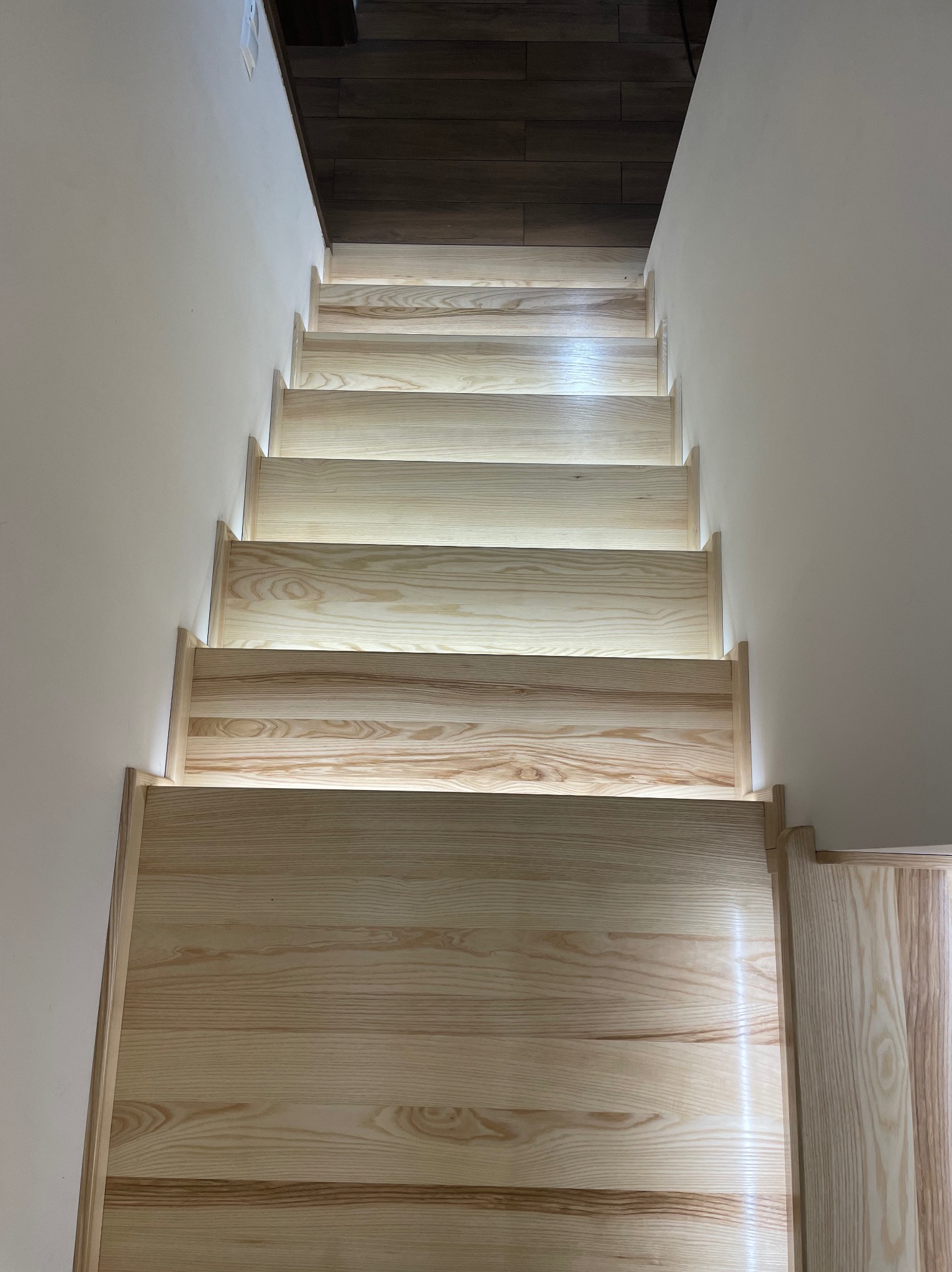 Jasne, drewniane schody z subtelnym podświetleniem LED wzdłuż stopni, prowadzące w górę. Minimalistyczny design i ciepłe oświetlenie akcentują naturalny rysunek drewna.