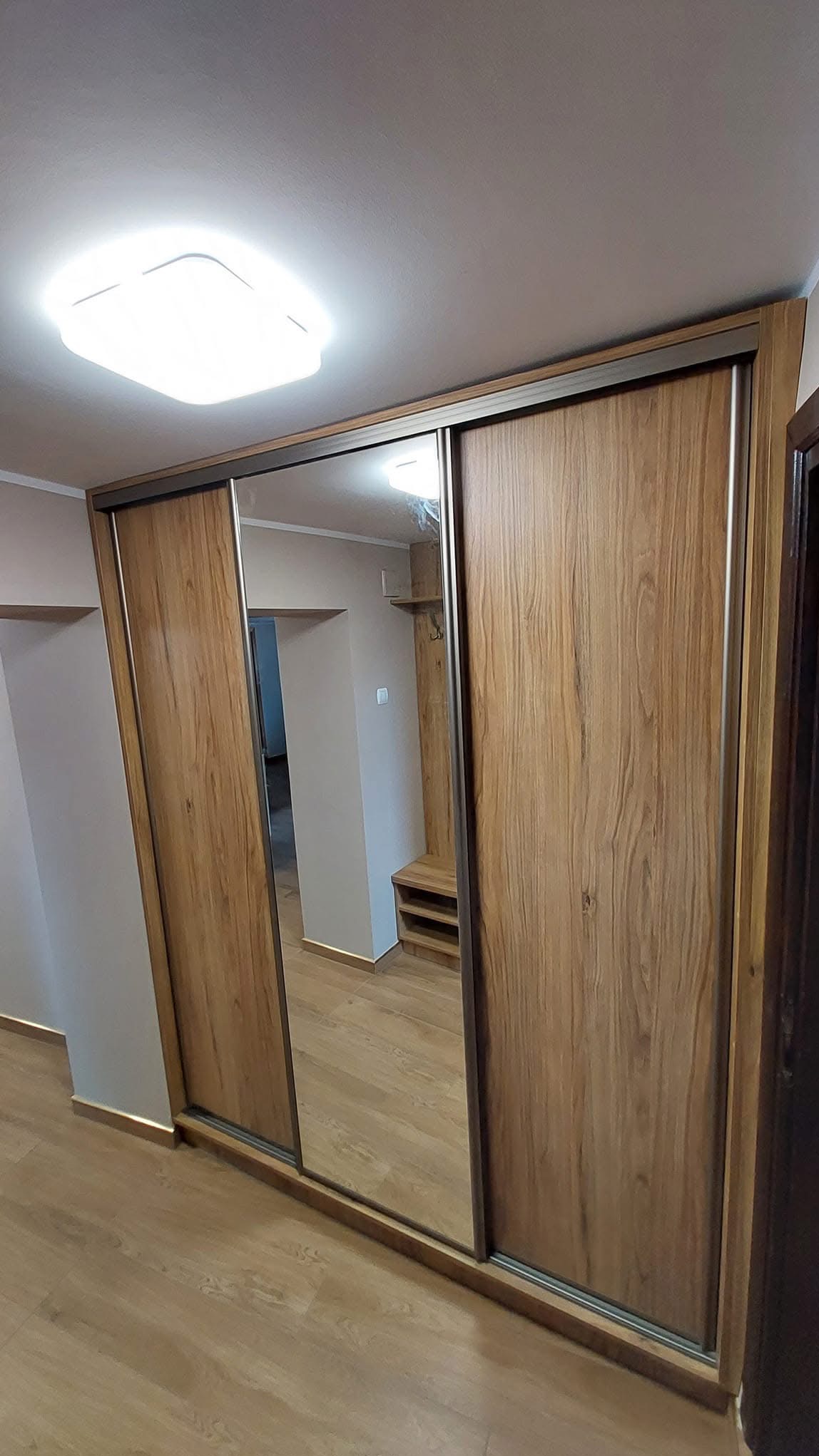 Szafa wnękowa z przesuwnymi drzwiami lustrzanymi, wykończona okleiną drewnopodobną, z widocznym fragmentem wnętrza i oświetleniem sufitowym.