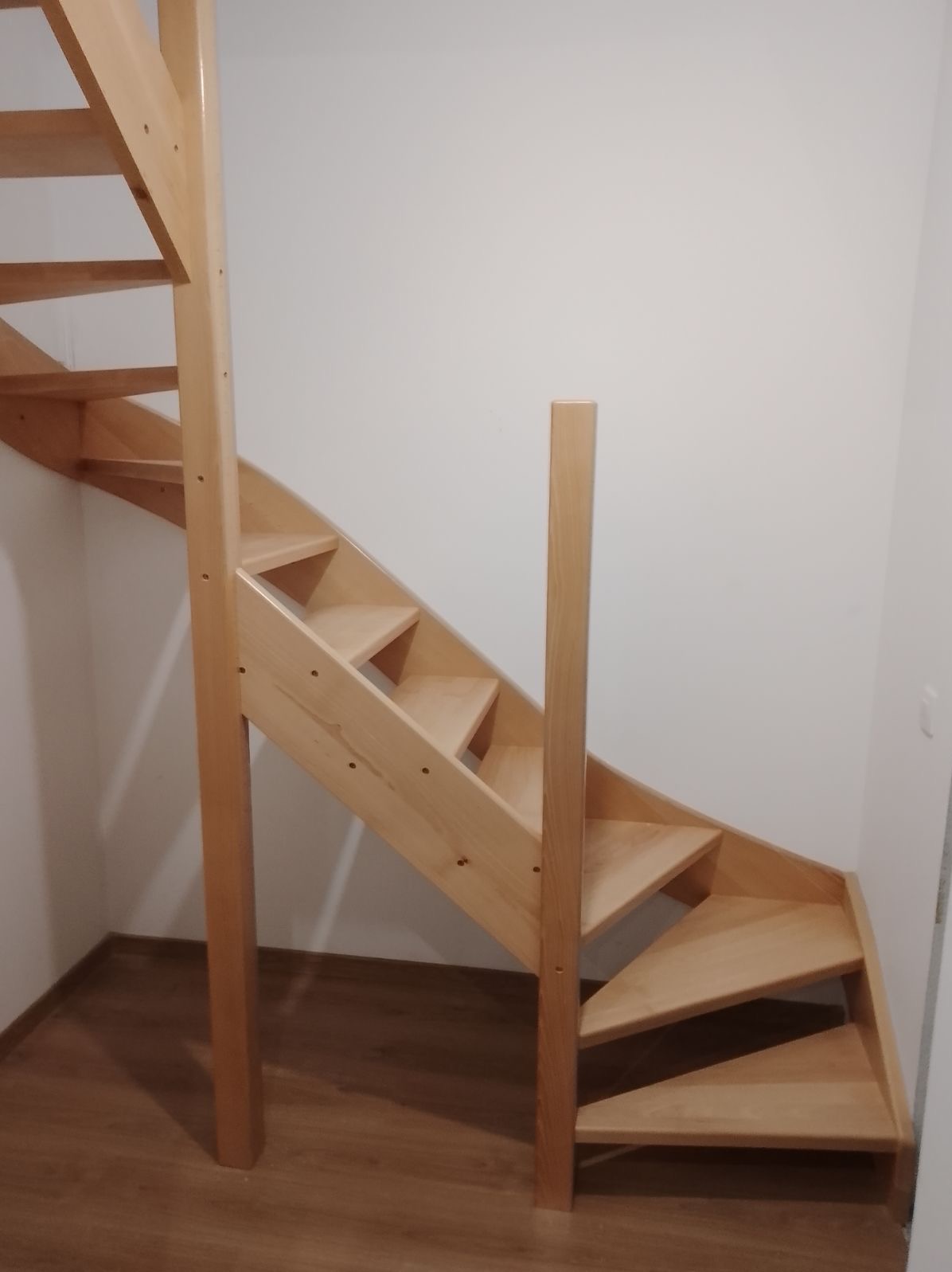 Jasne, drewniane schody z prostymi stopniami i pionowymi słupkami, zamontowane w narożniku pomieszczenia na panelach podłogowych.