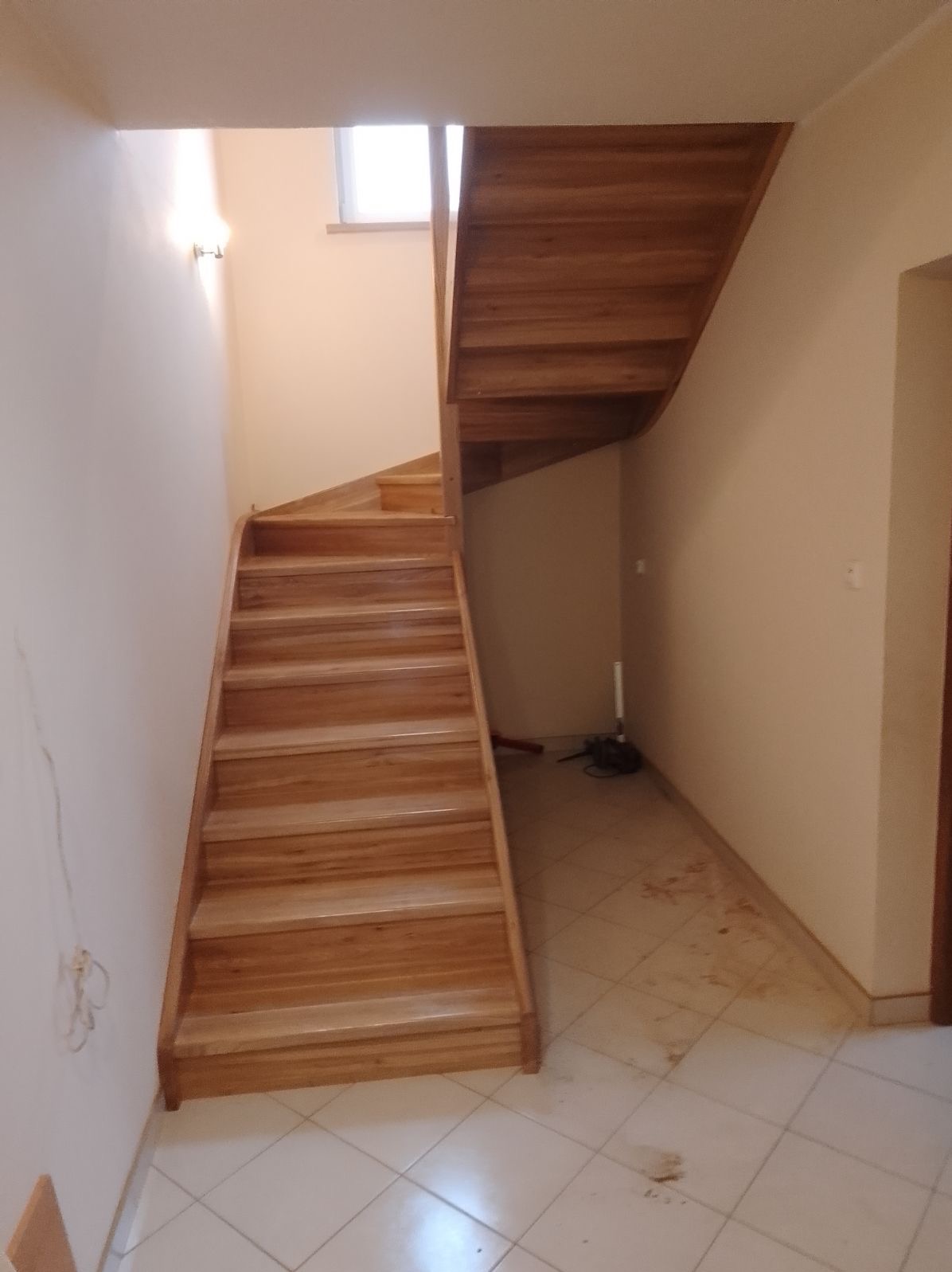 Jasne, drewniane schody z podstopnicami w jasnym wnętrzu. Widoczny fragment podłogi i ściany. Neutralne oświetlenie.