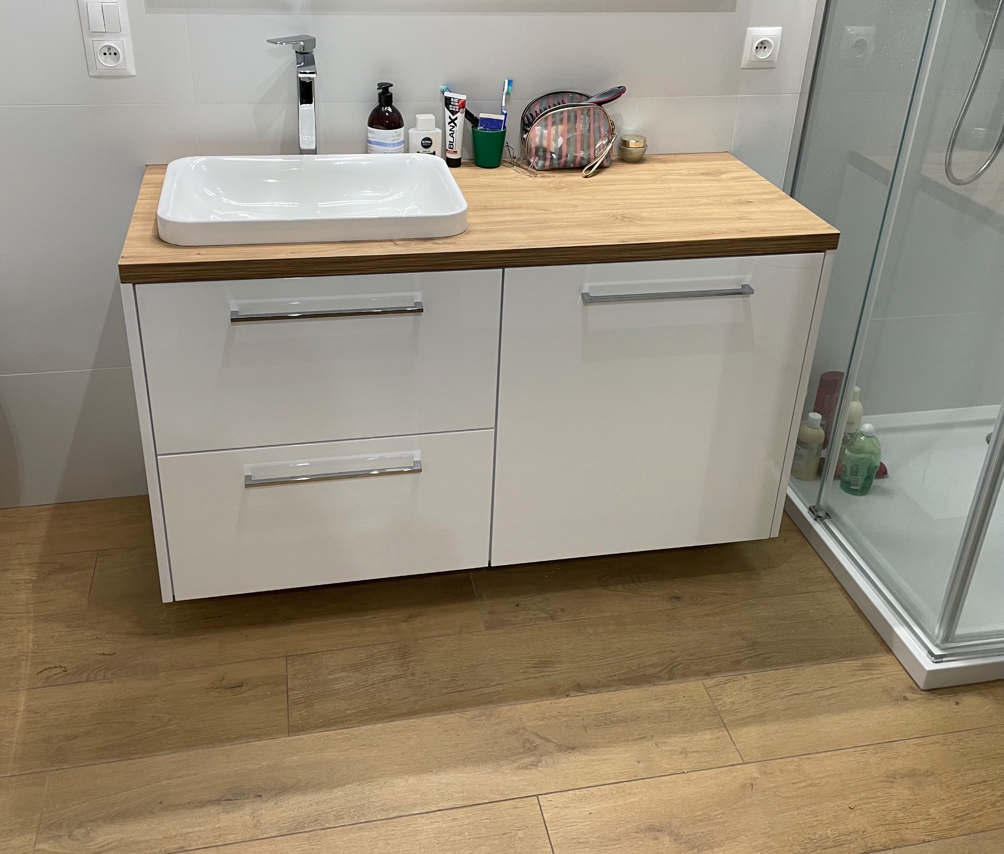 Nowoczesna szafka łazienkowa z blatem imitującym drewno i białą umywalką, obok kabina prysznicowa. Ujęcie z bliska, widoczne akcesoria kosmetyczne.