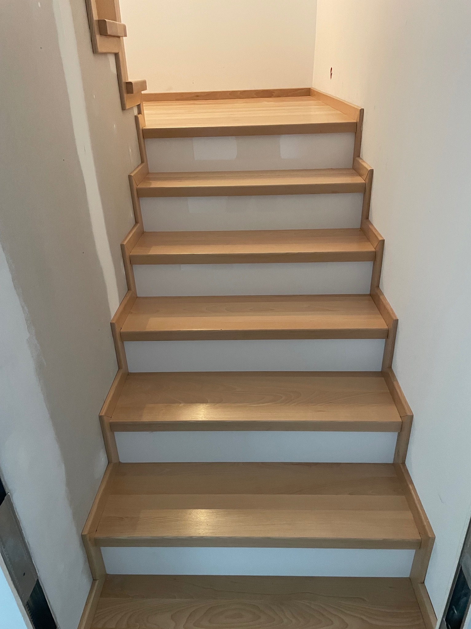 Jasne, drewniane schody z białymi podstopnicami, widok z dołu. Ściana po lewej w trakcie wykończenia, po prawej gładka, biała. Naturalne oświetlenie.