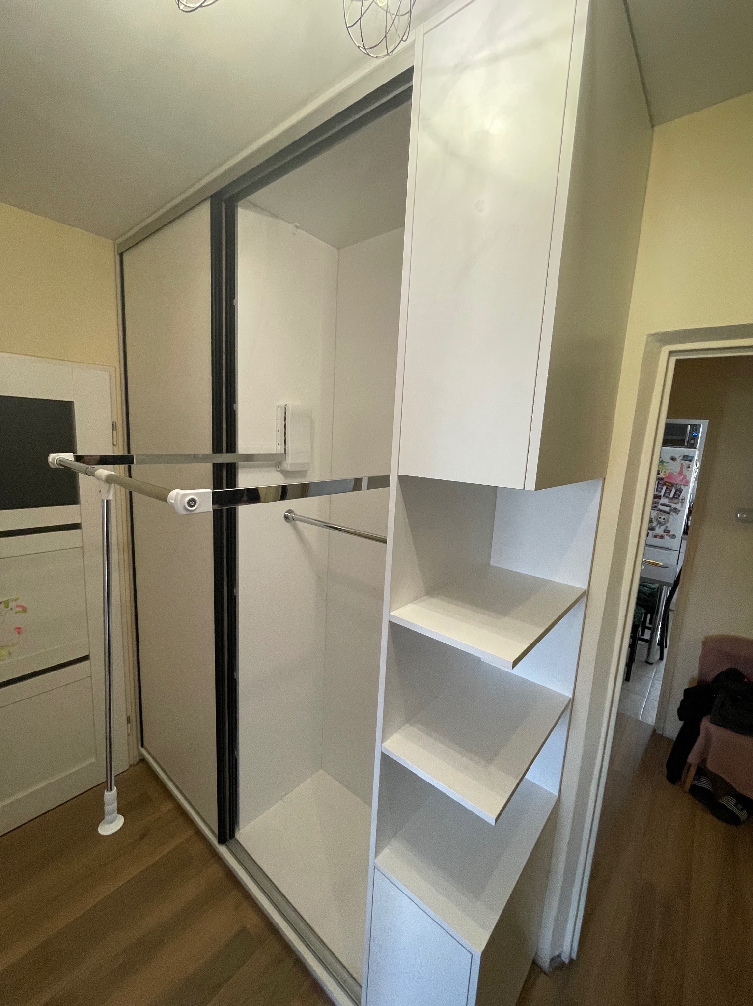 Biała szafa wnękowa z przesuwnymi drzwiami i półkami, widoczny system wieszaków. Ujęcie z perspektywy wnętrza, ukazujące detale wykończenia i funkcjonalność.