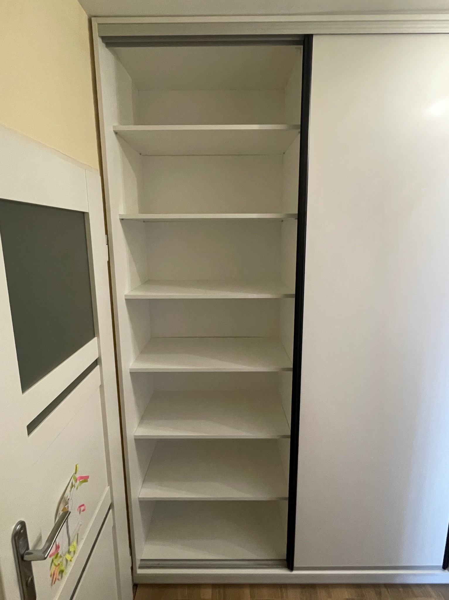 Biała szafa wnękowa z przesuwnymi drzwiami, z widocznymi półkami w środku, na tle jasnej ściany. Minimalistyczny design, widoczny fragment podłogi.