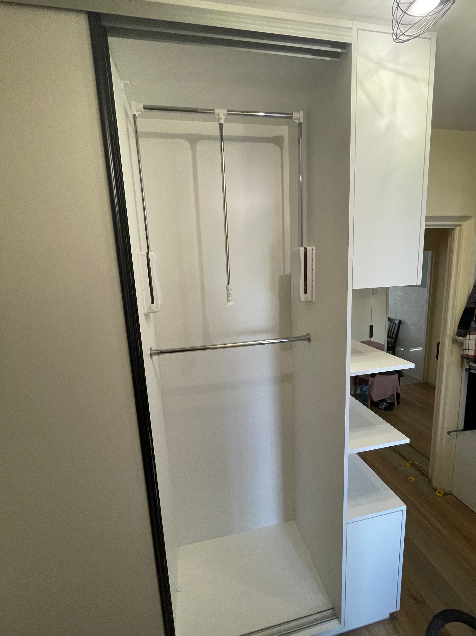 Wnętrze szafy wnękowej z białymi półkami i drążkami na ubrania. Widoczny system podnoszenia drążka. W tle inne pomieszczenie.