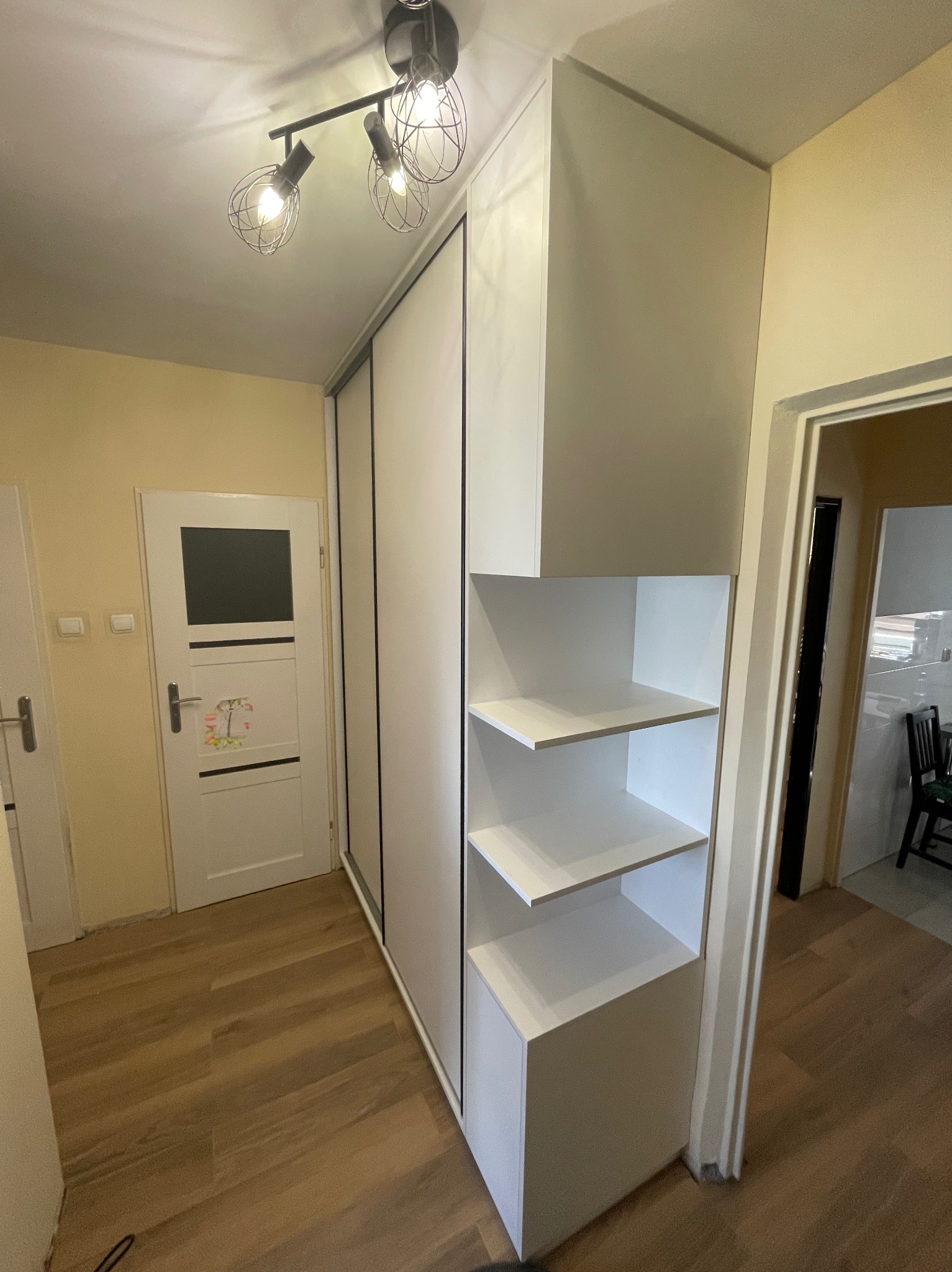 Biała szafa wnękowa z przesuwnymi drzwiami i otwartymi półkami, oświetlona lampą sufitową w korytarzu z drewnianą podłogą i białymi drzwiami.