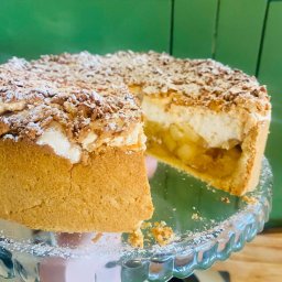 Karolina Zwierska - Krojone ciasto jabłkowe z kruszonką i cukrem pudrem na szklanej paterze, trzymane w dłoni na tle zielonej ściany. Widoczny przekrój z jabłkami i pianką.