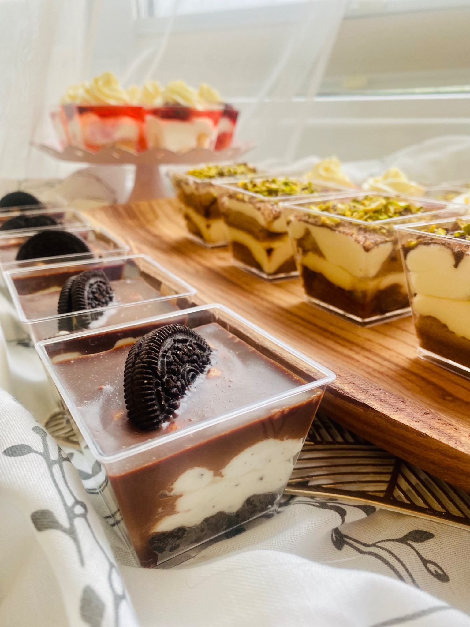 Deserki w przezroczystych pojemnikach: czekoladowe z Oreo, tiramisu z pistacjami i galaretka z owocami na drewnianej desce, w tle biała firanka.