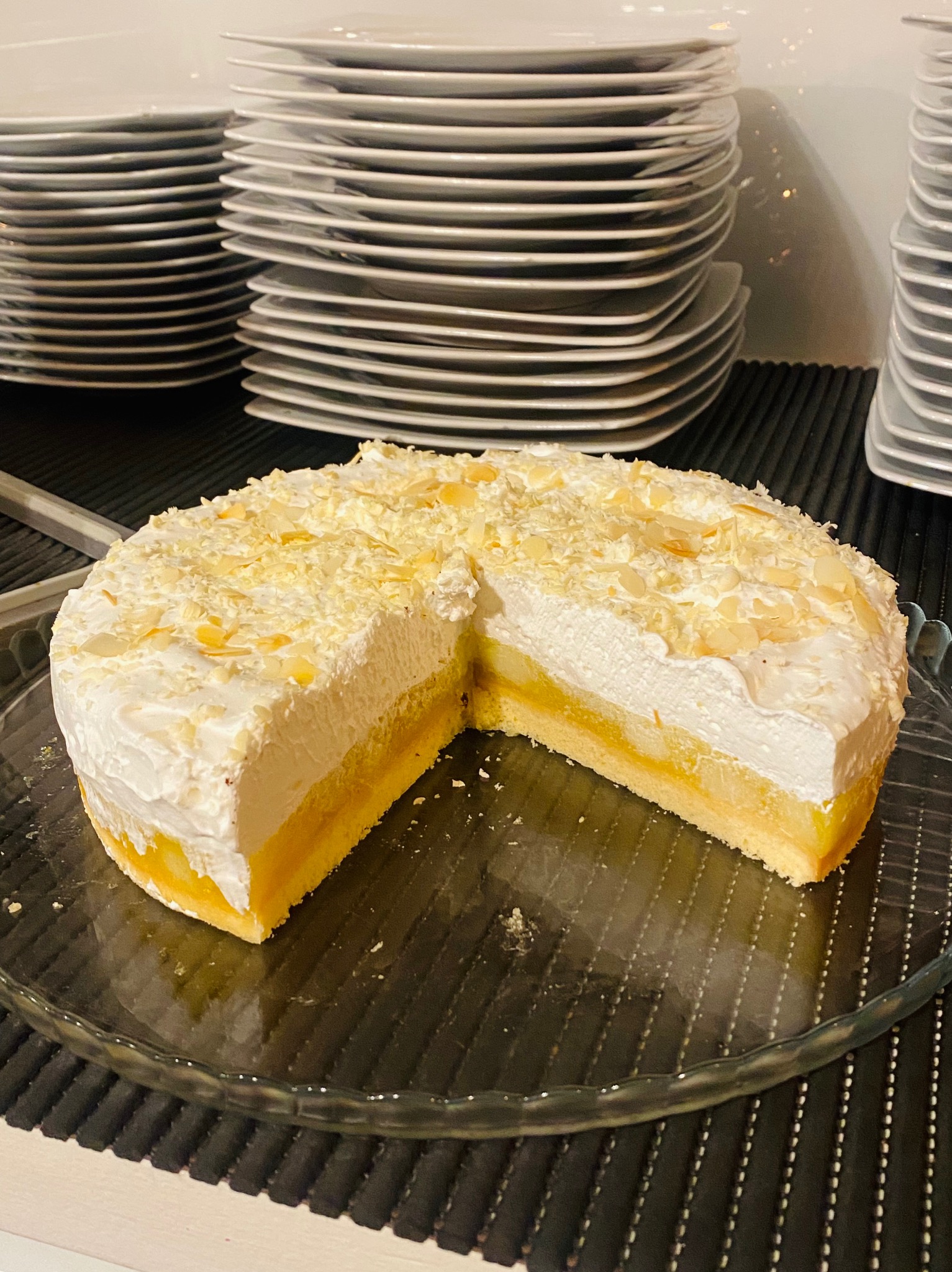 Kremowy tort z owocami i migdałami na szklanym talerzu, z wyciętym kawałkiem, w tle stosy białych talerzy.