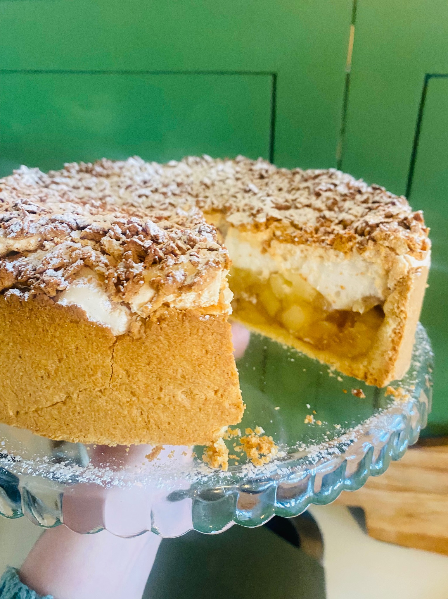 Krojone ciasto jabłkowe z kruszonką i cukrem pudrem na szklanej paterze, trzymane w dłoni na tle zielonej ściany. Widoczny przekrój z jabłkami i pianką.