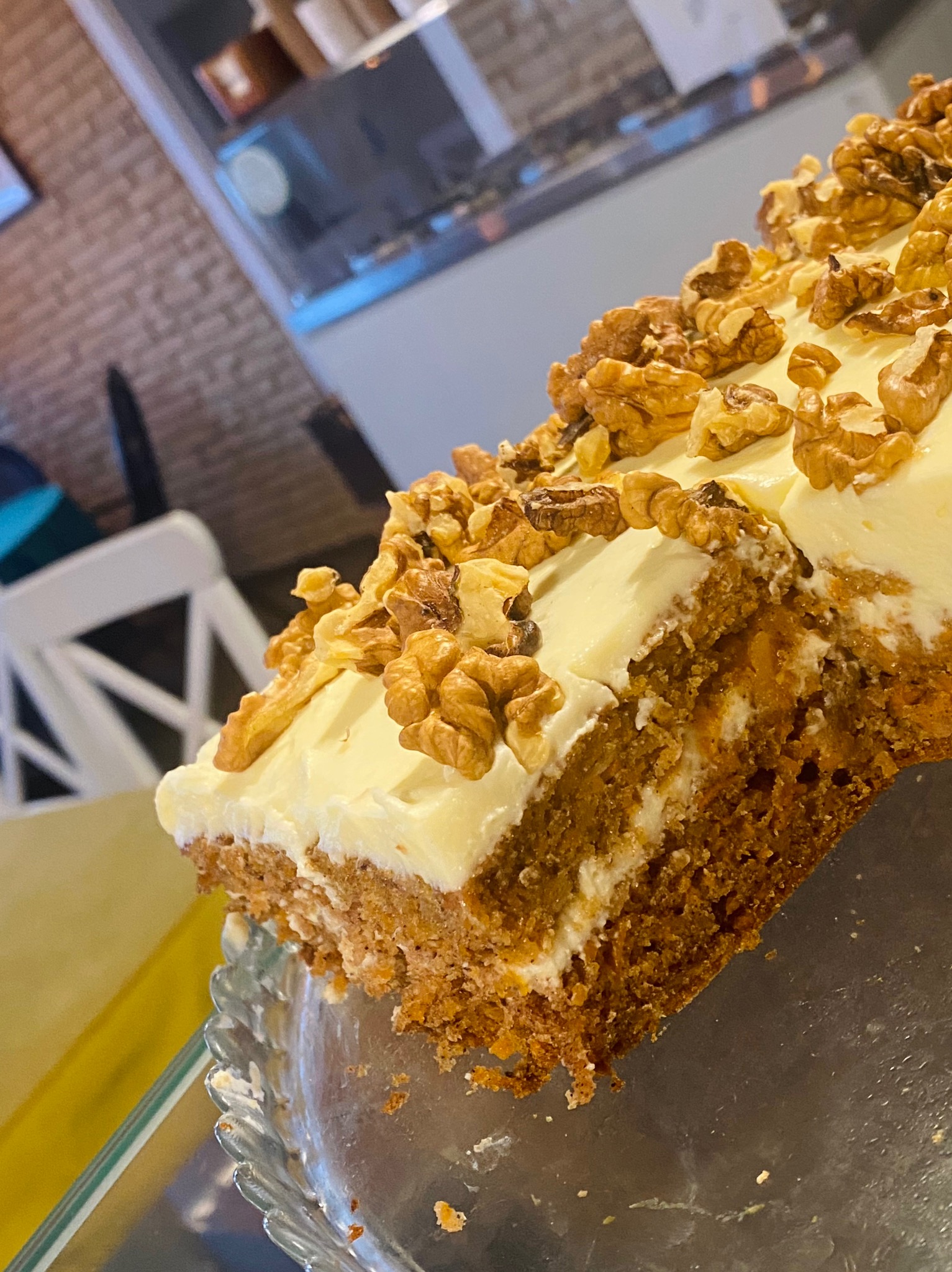 Kawałek ciasta marchewkowego z kremem i orzechami włoskimi, widoczny z bliska na szklanym talerzu, w tle wnętrze cukierni.